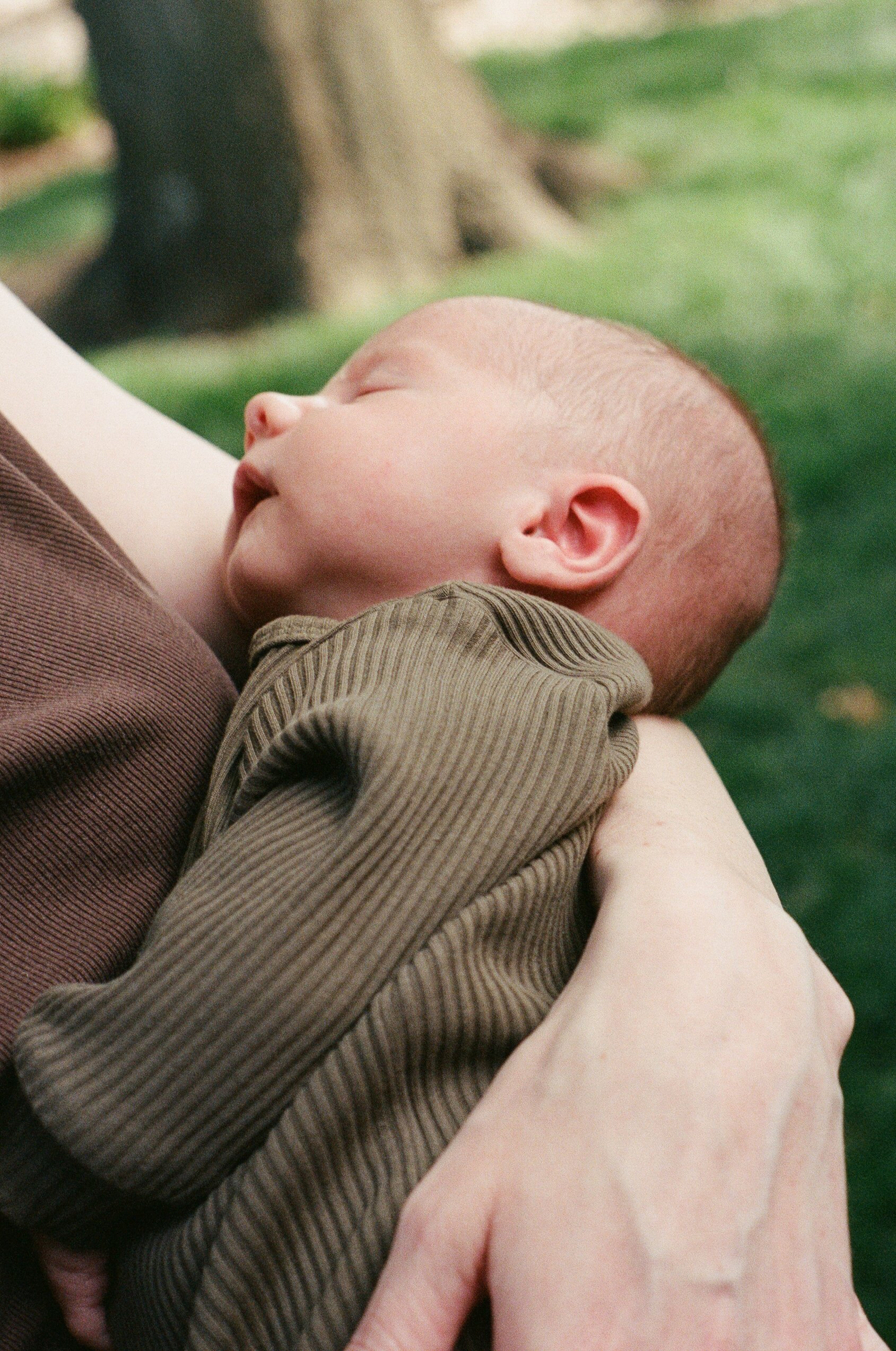 A young child with a shaved head sleeping peacefully in someone's arms, with trees and green grass in the background.
