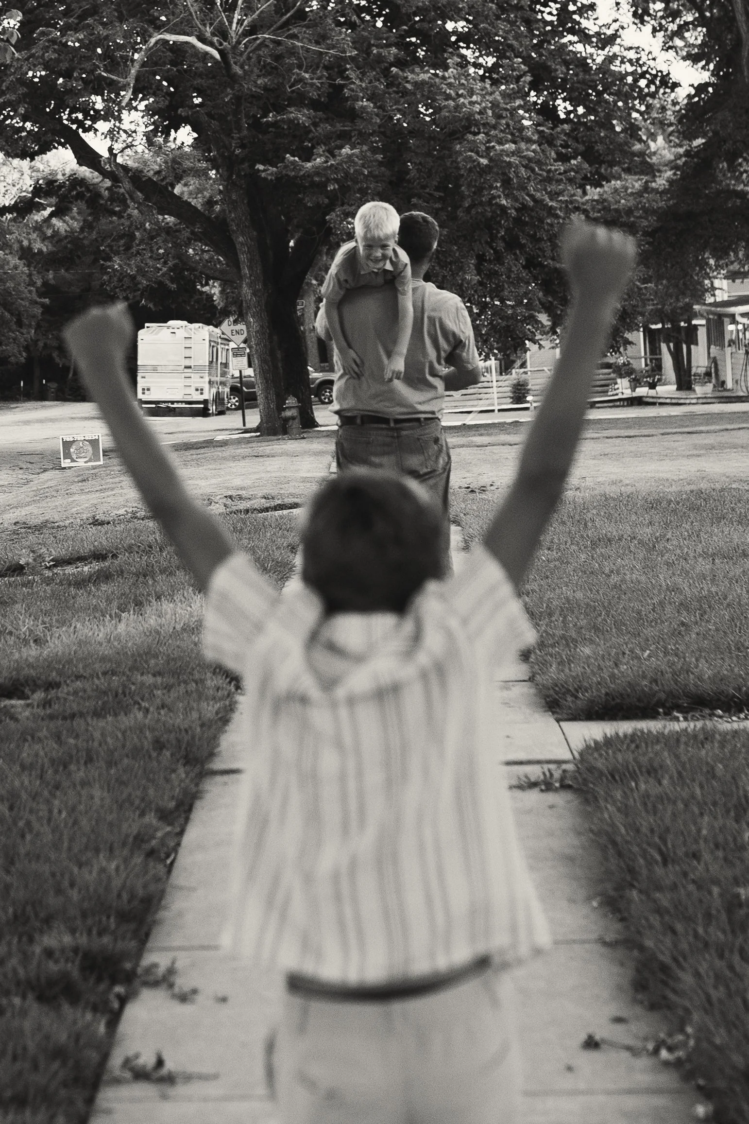 A boy with arms raised in the foreground facing away from the camera, with a small child held in an adult's arms in the background, who is smiling. The scene appears to be in a neighborhood or park with trees and houses.