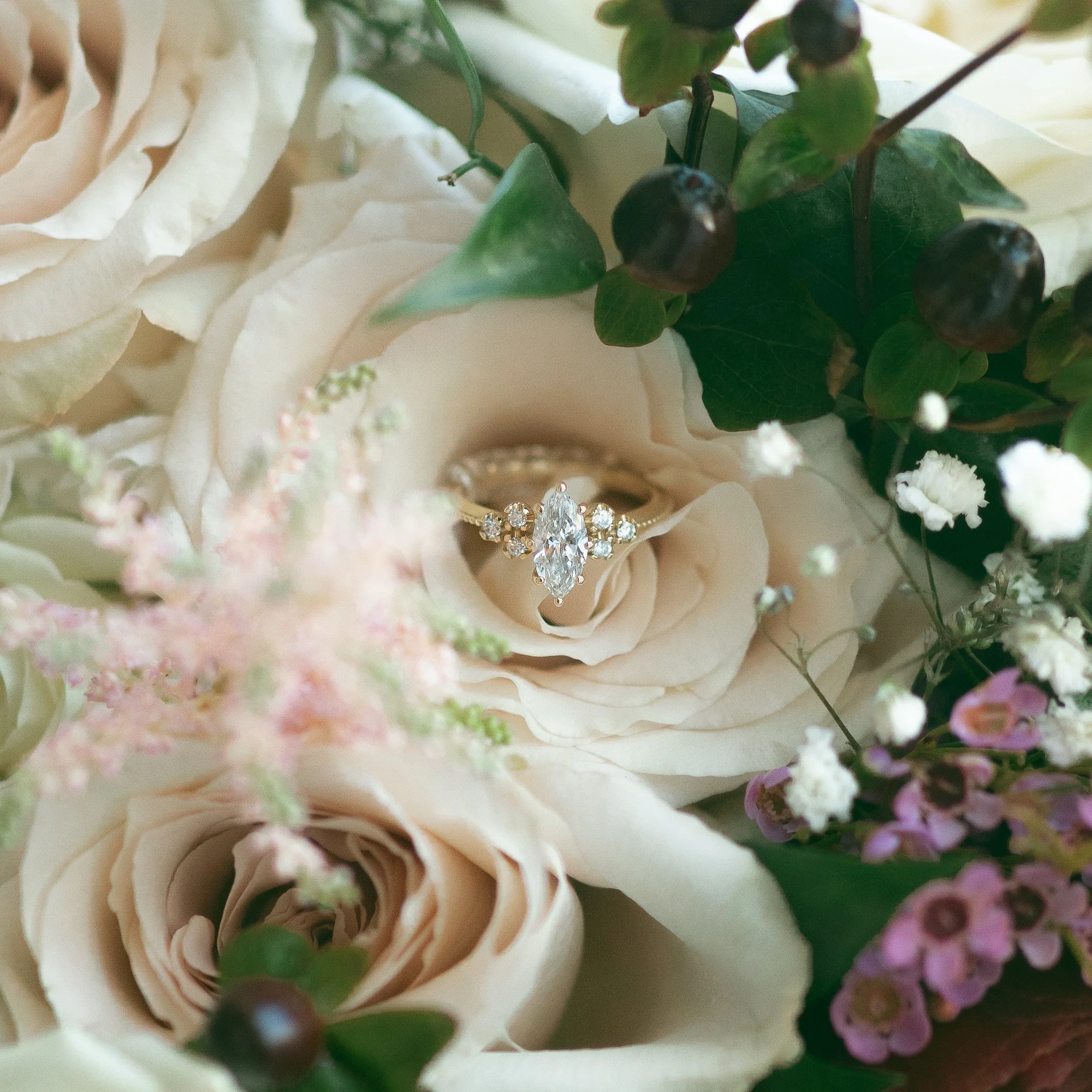 A diamond engagement ring resting inside a cream-colored rose surrounded by various flowers and green leaves.