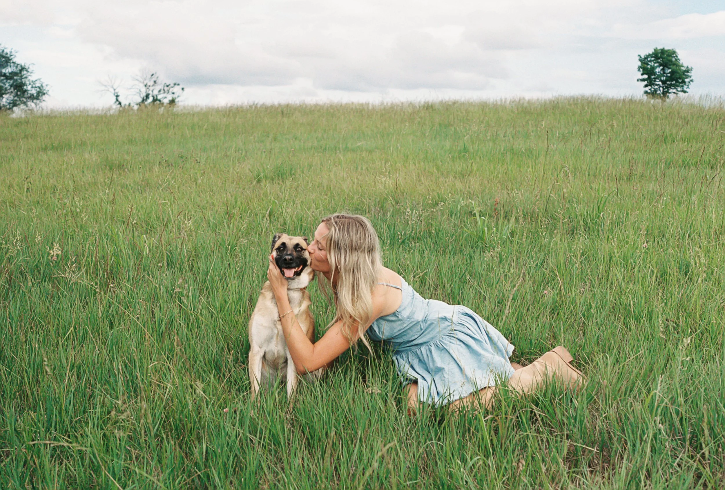 A woman with long blonde hair lying on her side in a green grassy field, holding and kissing a large tan and black dog with a happy expression, with a cloudy sky and distant trees in the background.