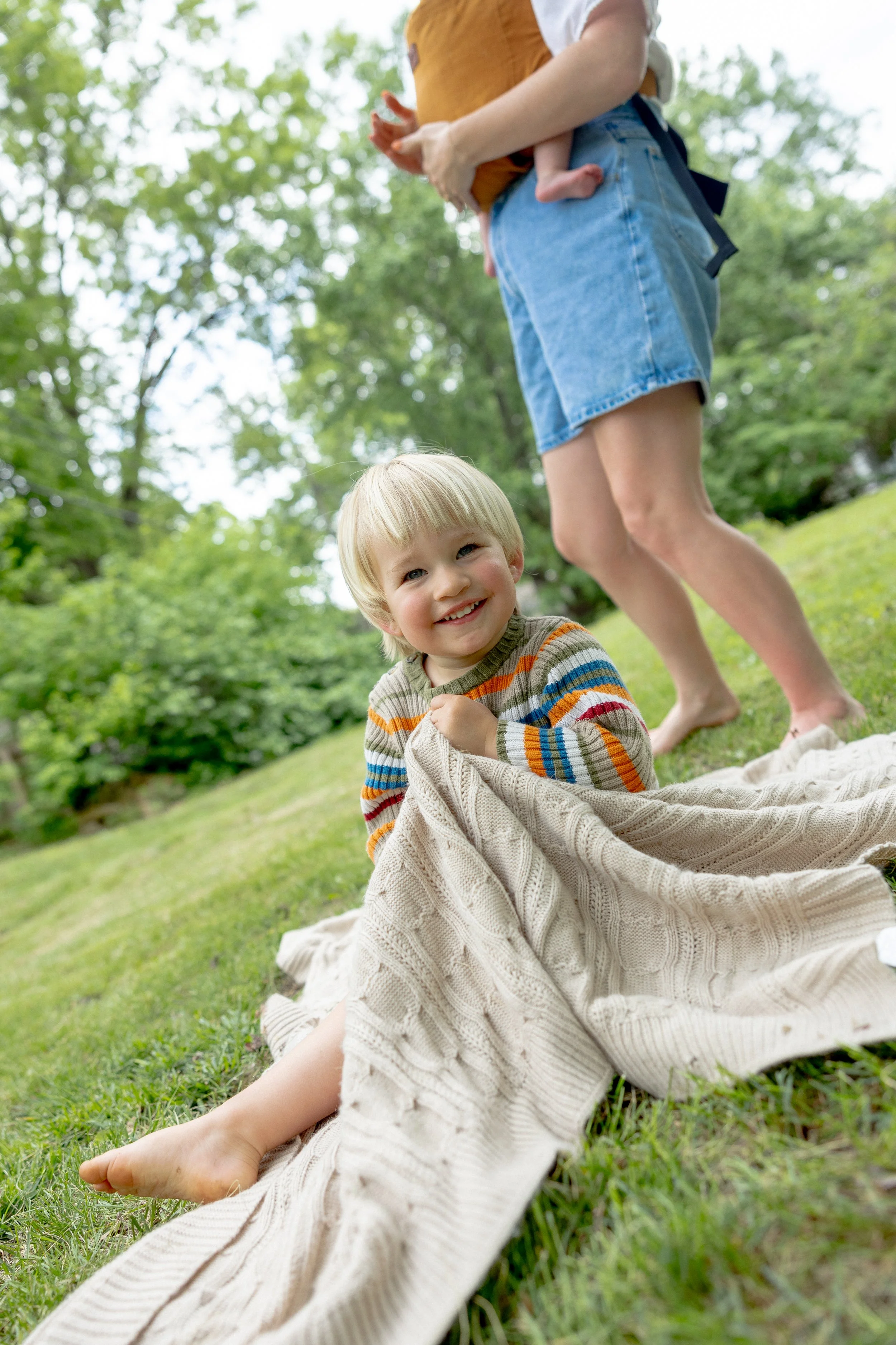 A smiling young boy with blonde hair is lying on the grass in a park, wrapped in a large, beige knitted blanket. An adult, wearing denim shorts and a brown top, is standing nearby holding a small child. The background features lush green trees and a 