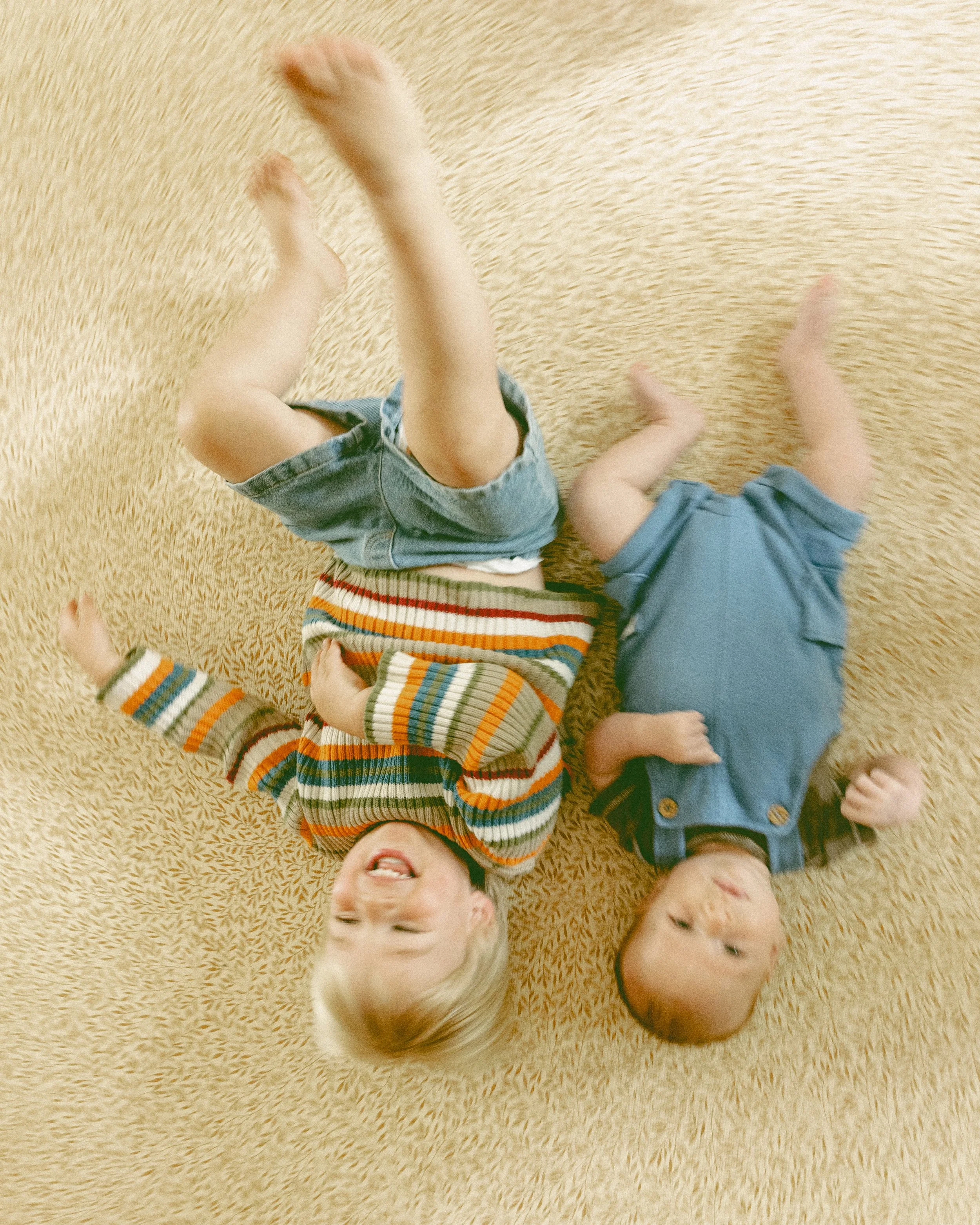 Two young children lying on a beige carpet, smiling and playing. One child has blonde hair, wearing a striped sweater and denim shorts. The other child is bald, wearing a blue shirt.