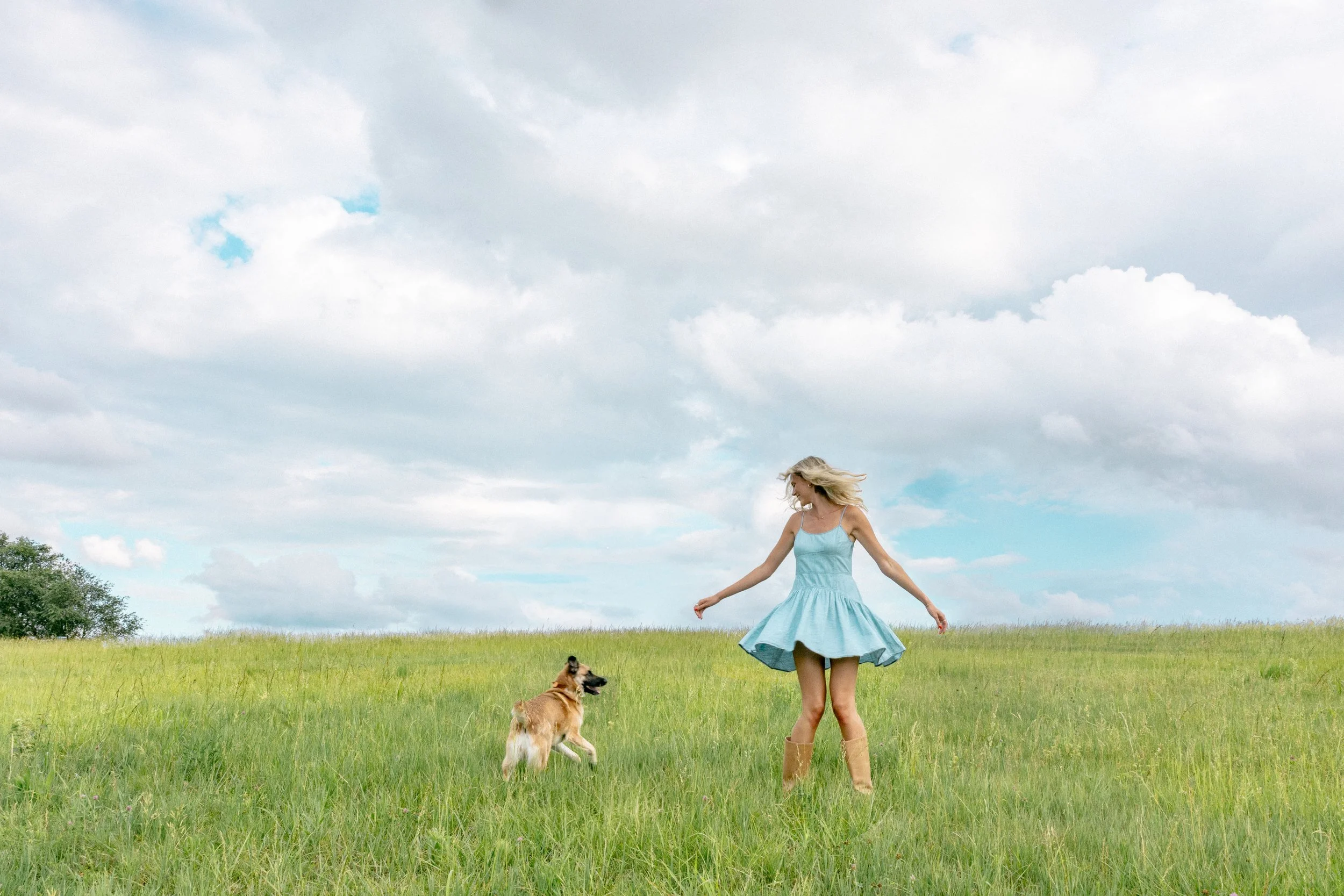 A woman in a blue dress and brown boots is dancing in a green field with a dog, under a cloudy sky.