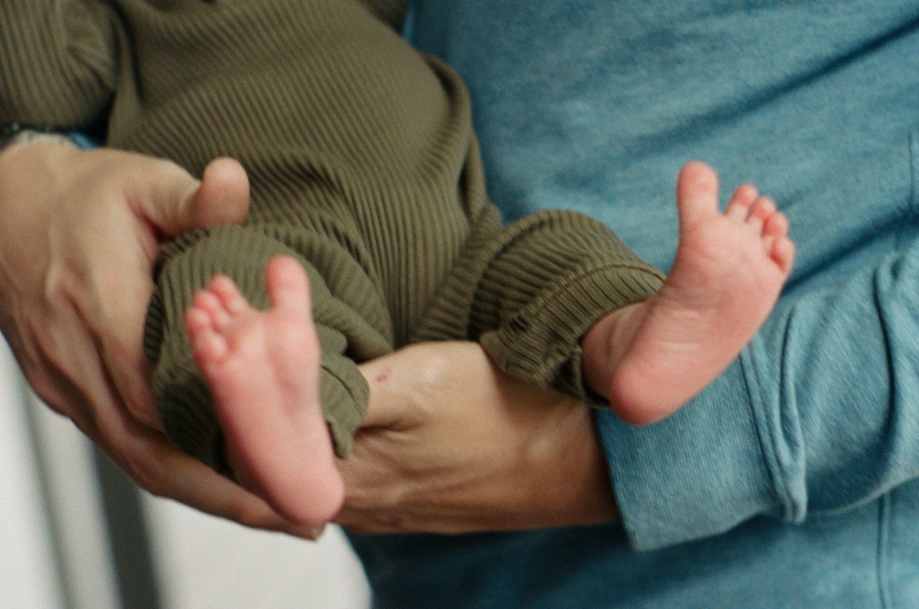 A person holding a newborn baby's legs and feet, with the baby wearing brown pants and lying against an adult wearing blue jeans.