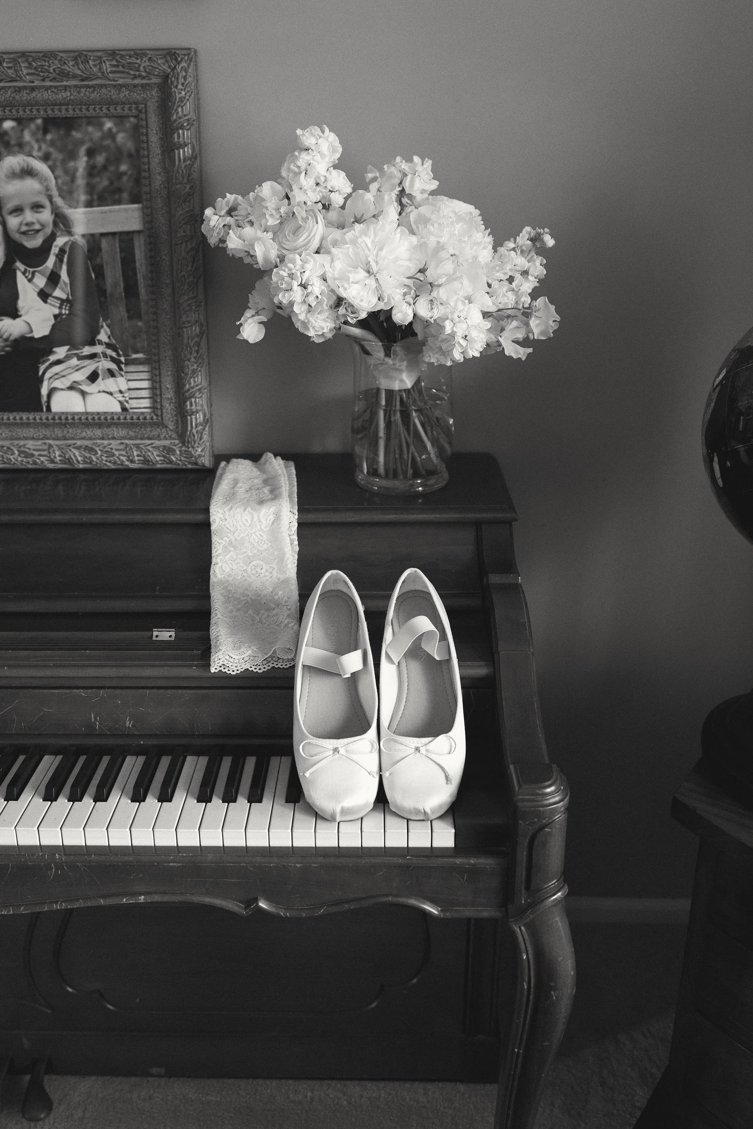 A pair of white ballet flats with bows on the toes placed on the keys of a piano, with a framed photograph of a smiling girl, a vase with white flowers, and lace fabric on the piano. A helmet and part of a mirror are visible in the background.