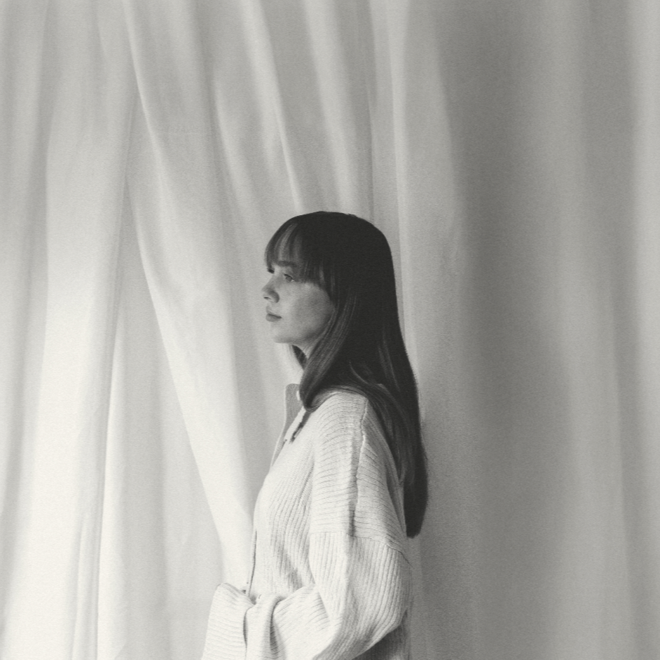 A woman standing next to light-colored curtains, looking out with a calm expression.