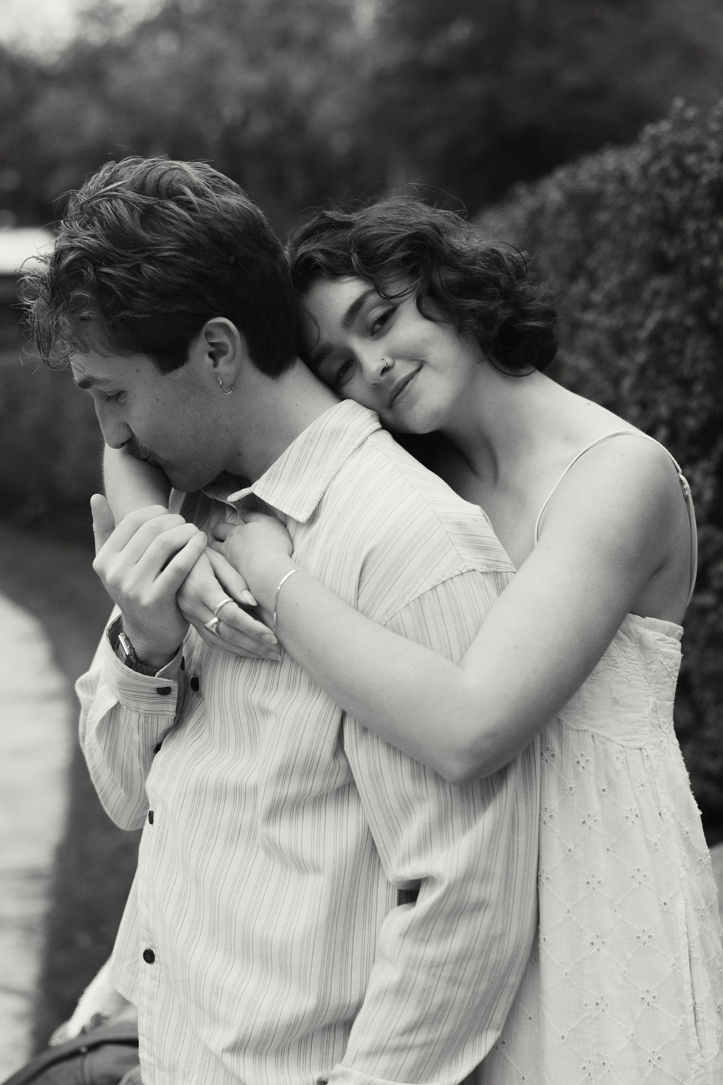 A black-and-white photo of a young couple embracing outdoors, with the woman smiling and resting her head on the man's shoulder.