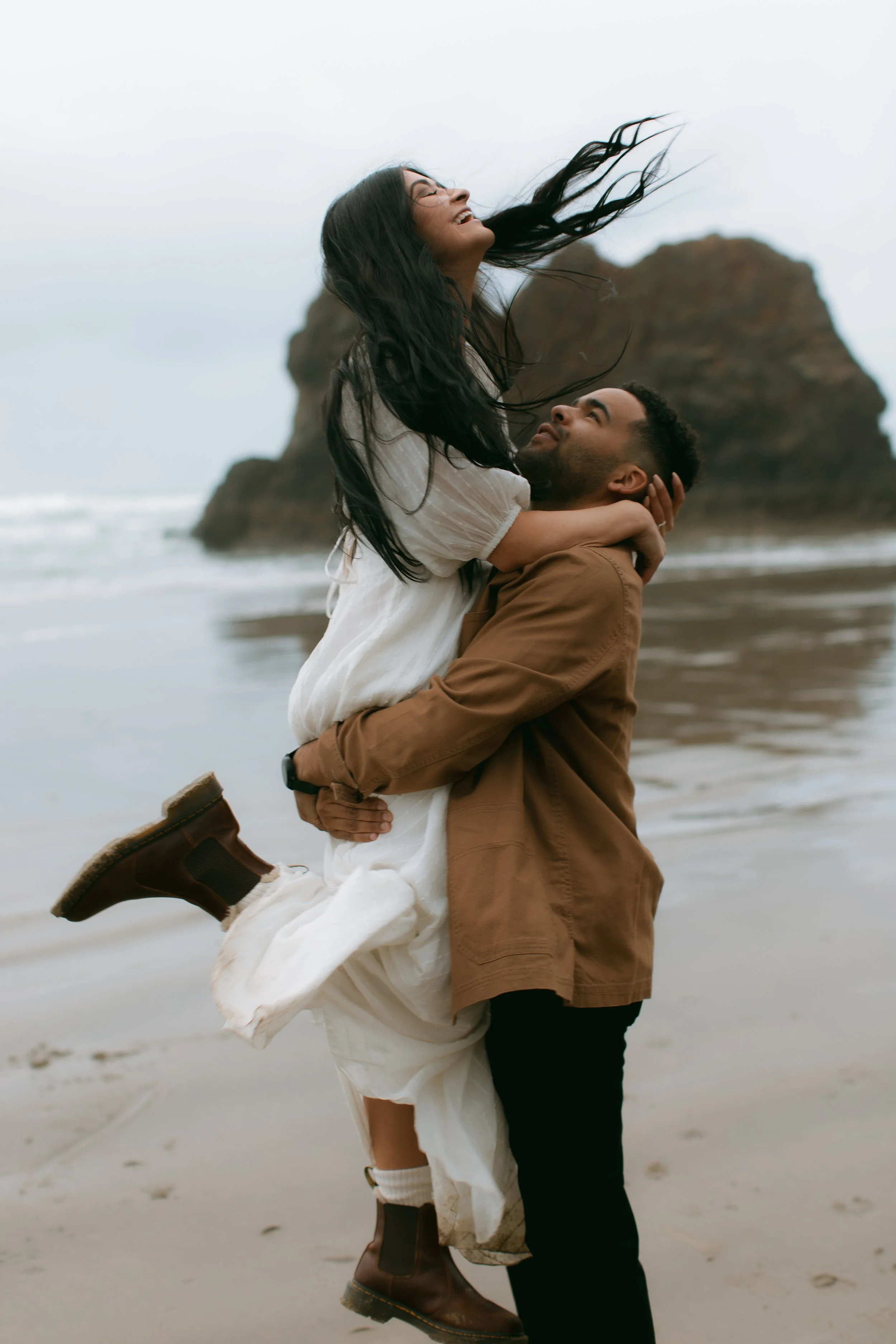 A couple at the beach, with the man lifting the woman, she has her long dark hair flowing, and they are smiling. The background shows ocean waves and large rock formations.