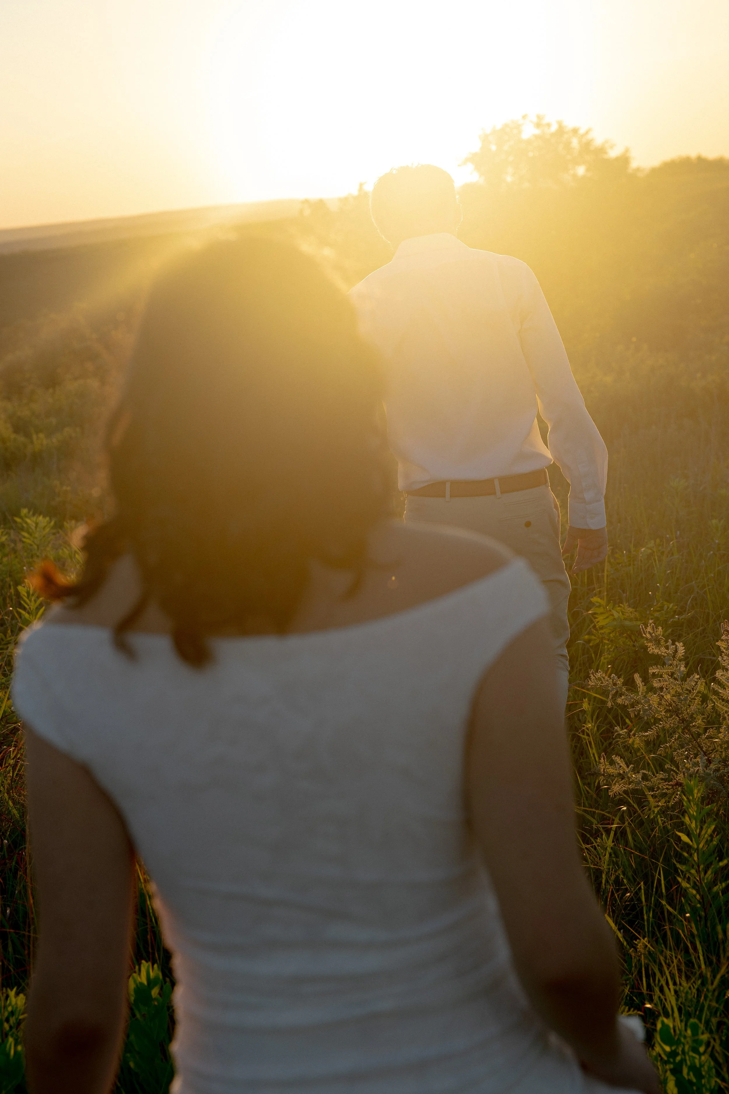 A person with curly hair wearing a white sleeveless top, standing outdoors among green plants at sunset or sunrise.