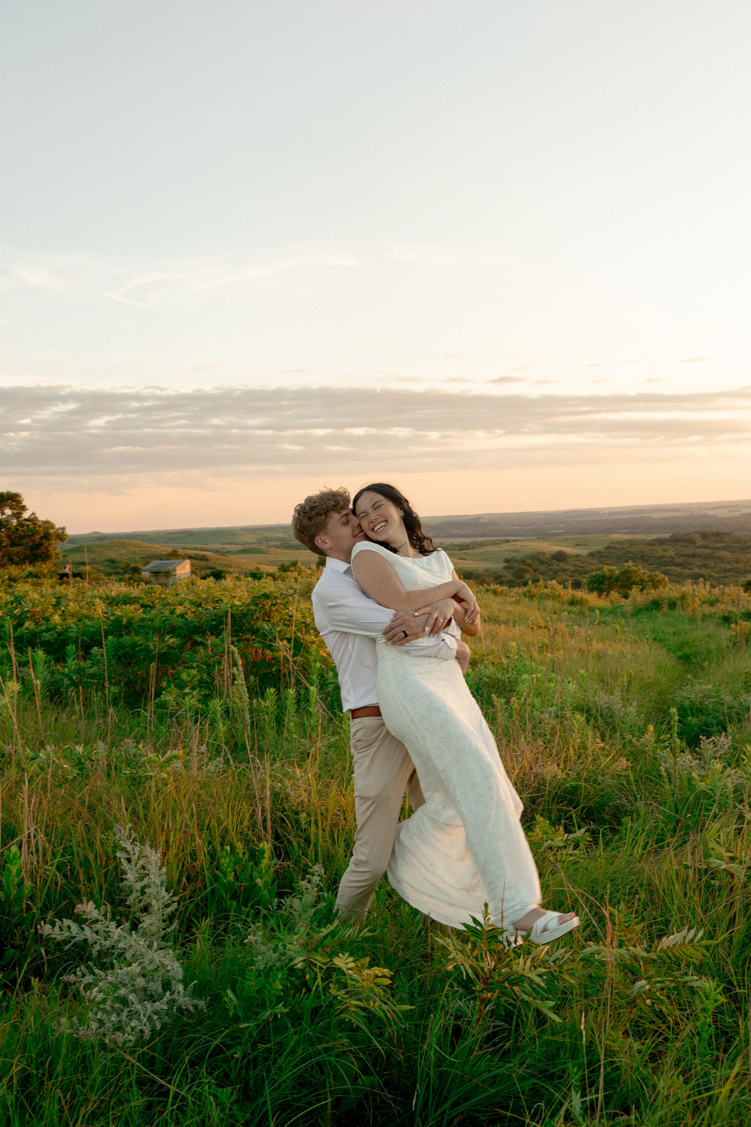 A couple happily embracing in a field of green plants at sunset, with rolling hills and a few scattered buildings in the background.