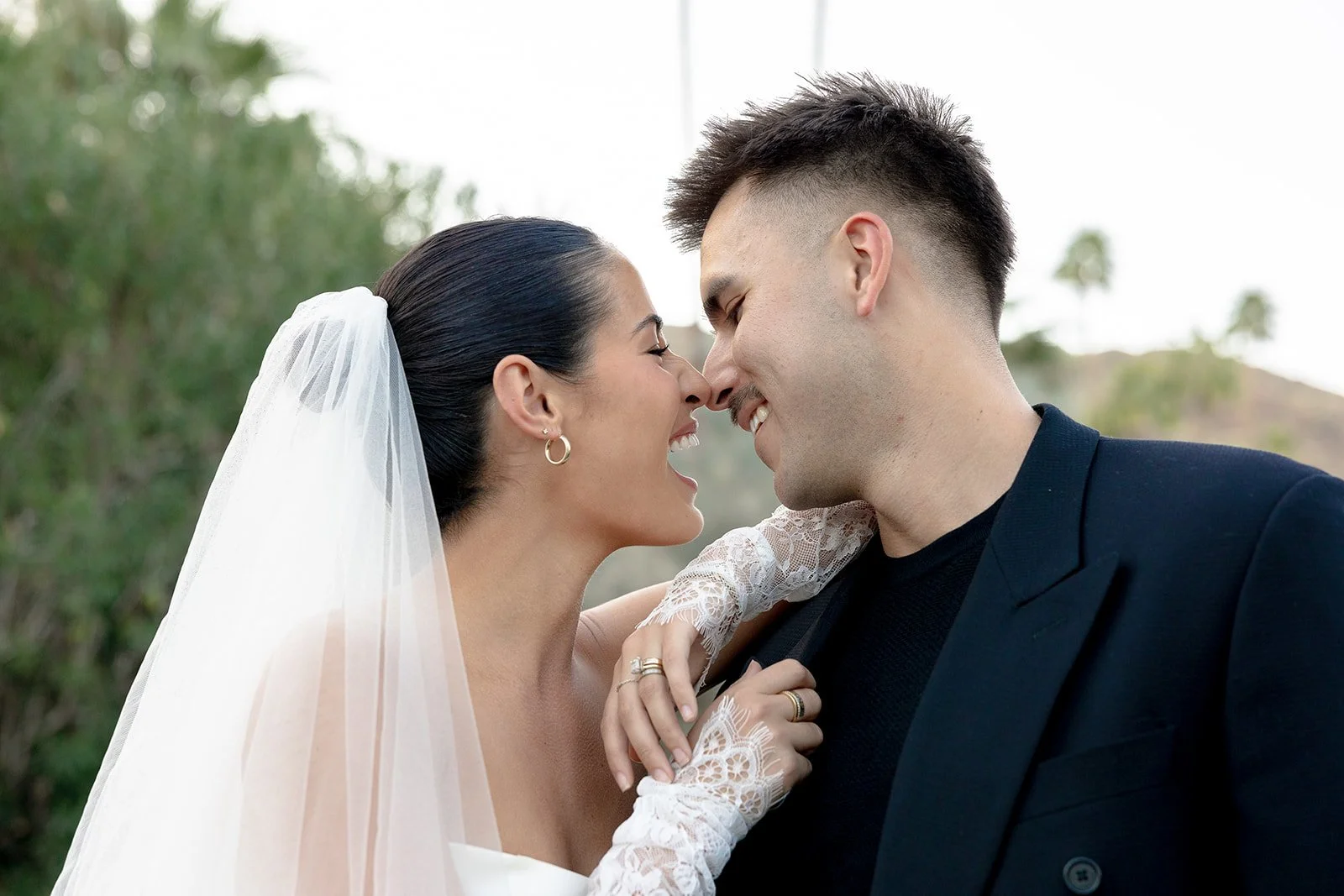 A bride and groom with foreheads touching, smiling, outdoors with trees and mountains in the background during daytime.