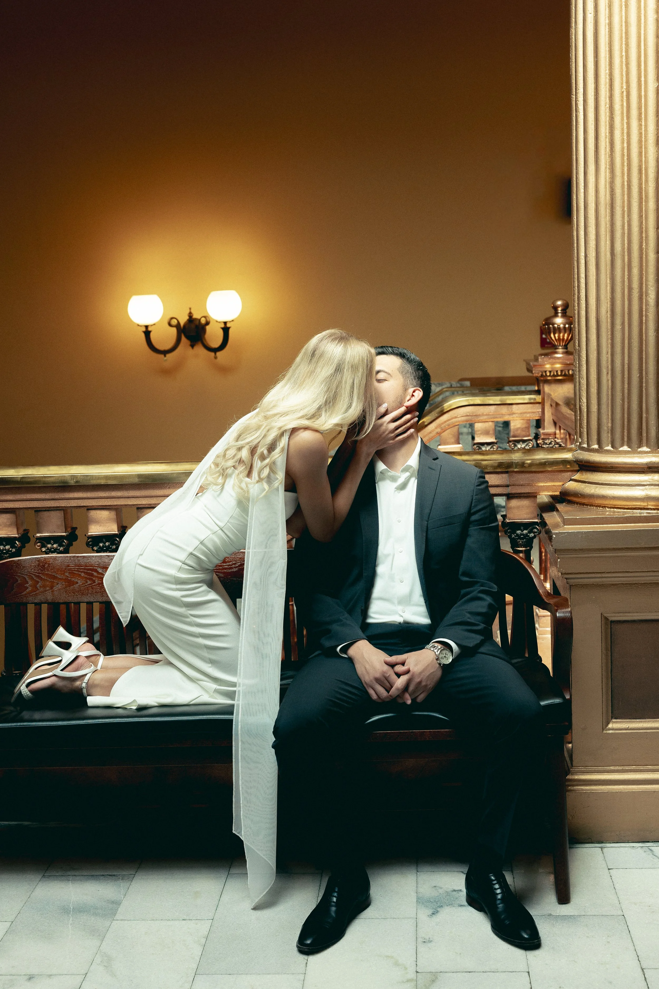 A woman in a white dress and heels kneeling on a bench, kissing a seated man in a tuxedo in a warmly lit interior with columns and wall sconces.