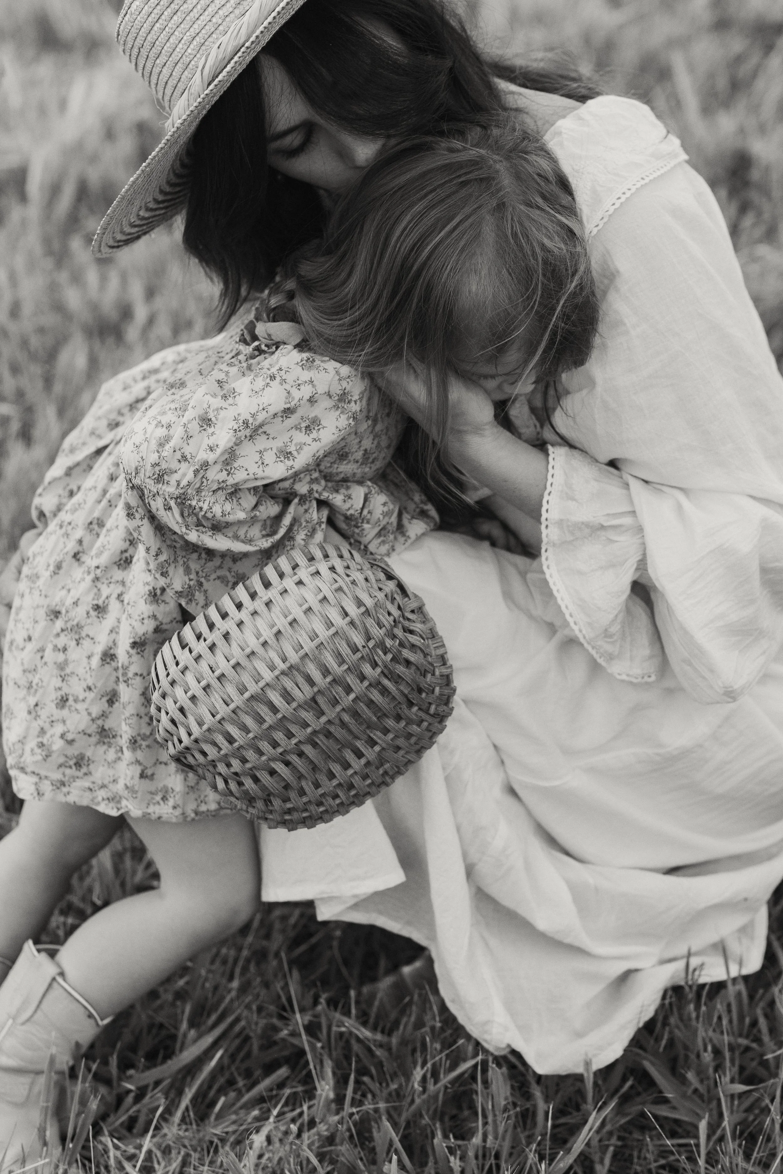 A woman and a young girl embracing each other outdoors in a grassy field, with the woman wearing a wide-brimmed hat and long sleeve dress, and the girl holding a round woven basket, both sharing a close, comforting hug.