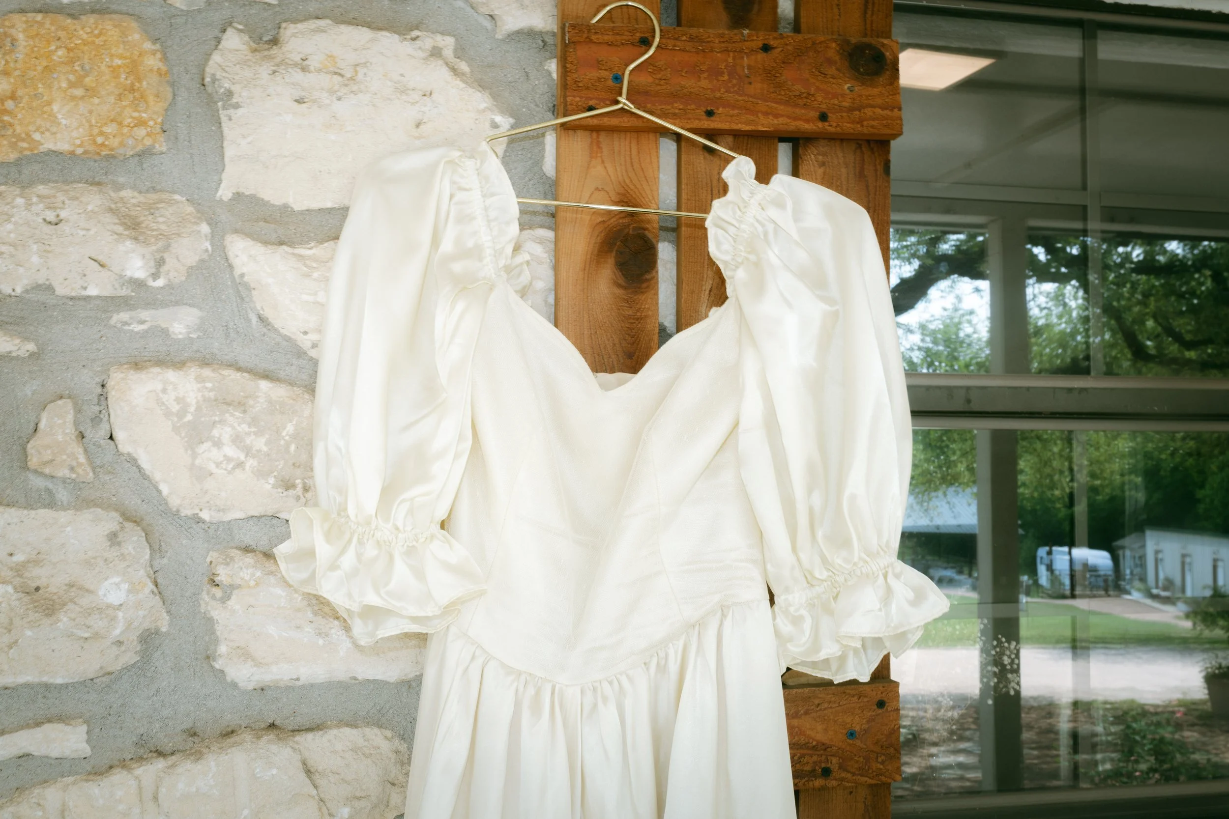 A white dress with puffed sleeves hanging on a wooden hanger against a stone wall with a window in the background.