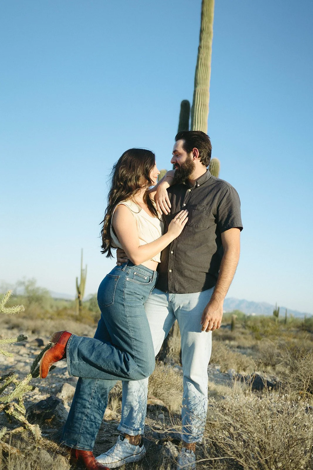 A man and woman in a desert landscape with tall cacti, standing close and looking at each other.