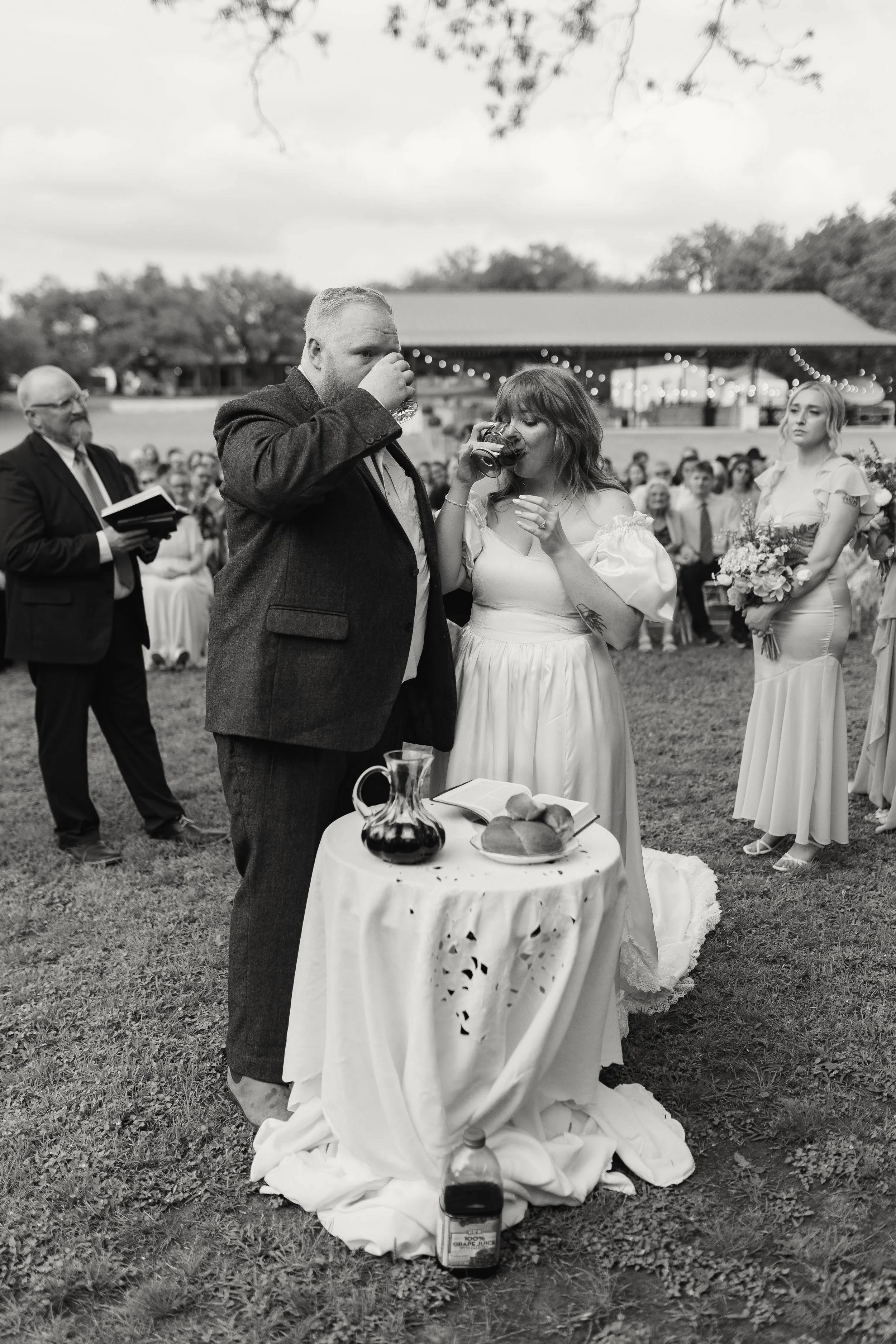 A black-and-white photo of a wedding ceremony outdoors with a bride and groom drinking from glasses. The bride is in a white dress holding a flower bouquet, and the groom in a dark suit. Guests are seated in the background under a pavilion decorated 