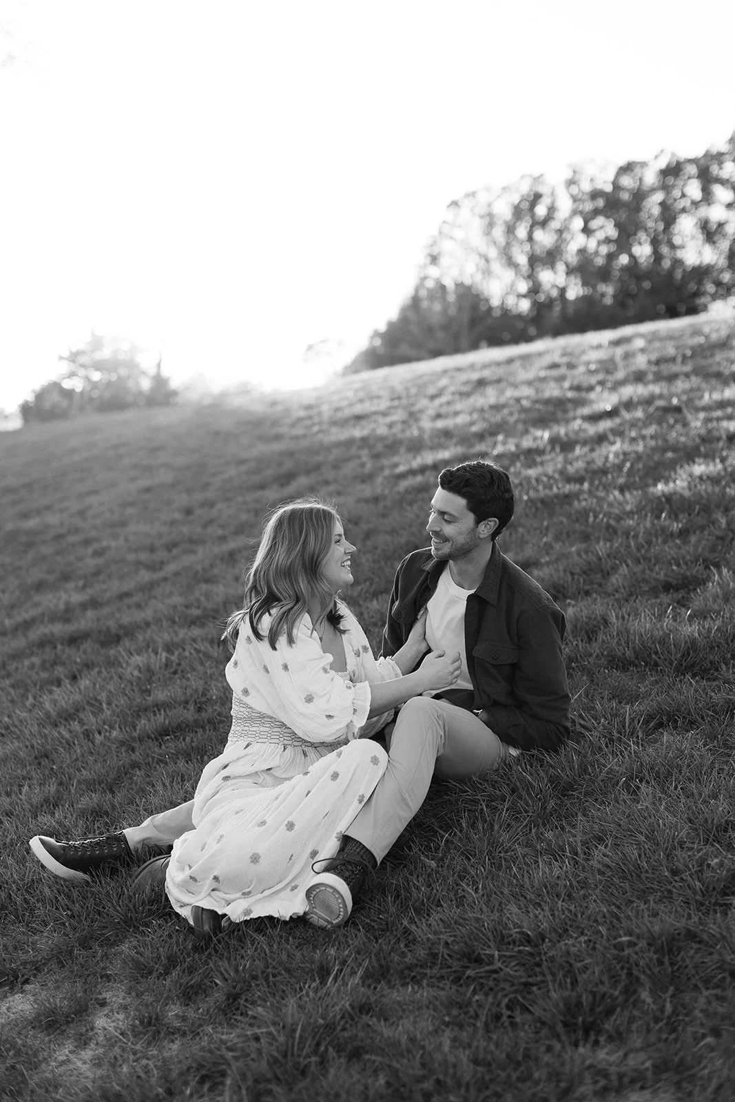 A couple sitting on the grass in a park, looking at each other and smiling, with trees in the background, captured in black and white.
