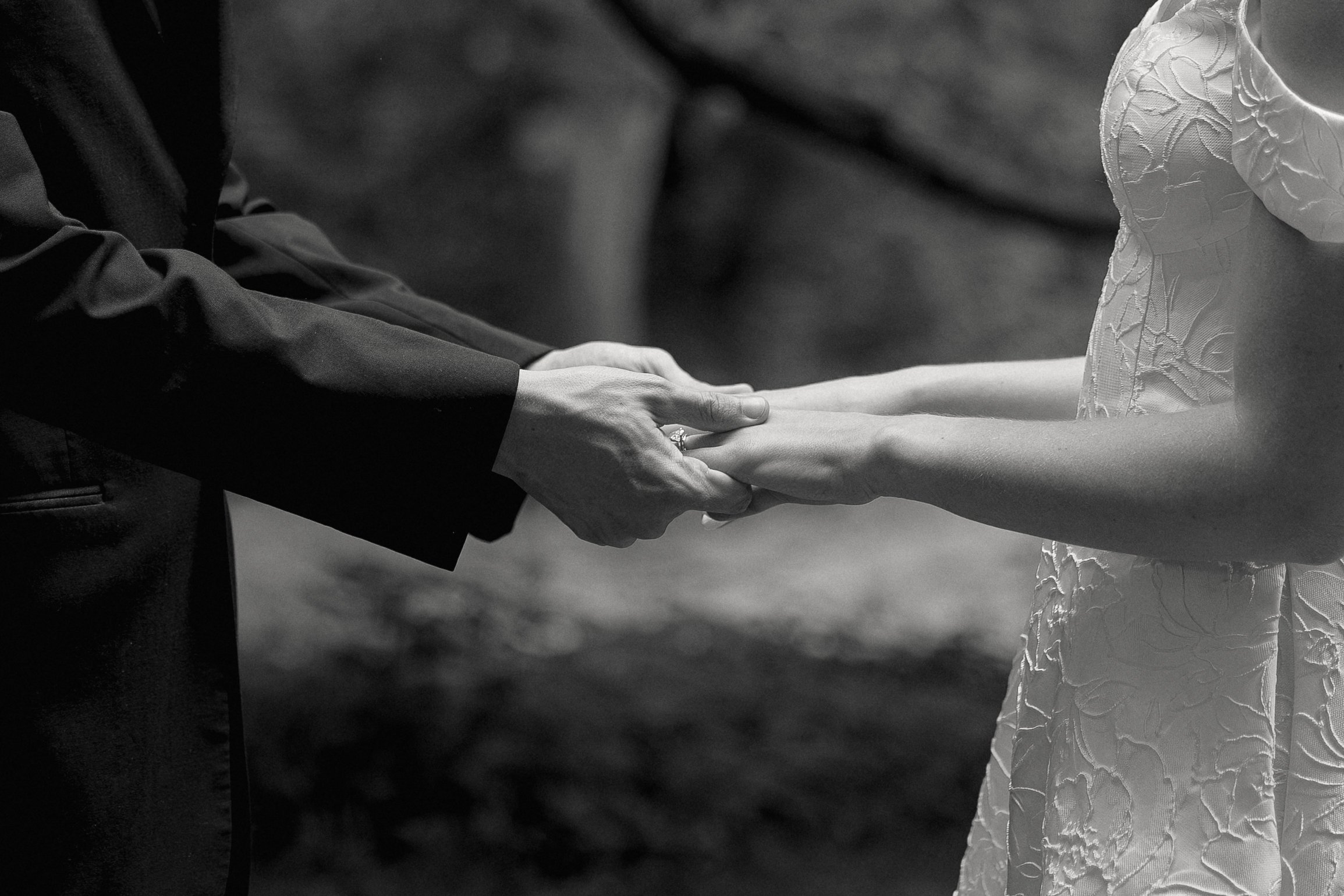 Black-and-white photo of a couple holding hands during a wedding ceremony, with the groom wearing a suit and the bride wearing a dress with embroidery.