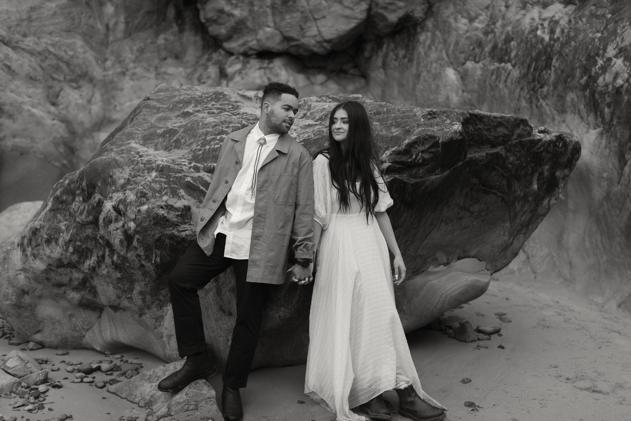 A young man and woman holding hands next to a large rock on a beach, with rocky cliffs in the background, in black and white.