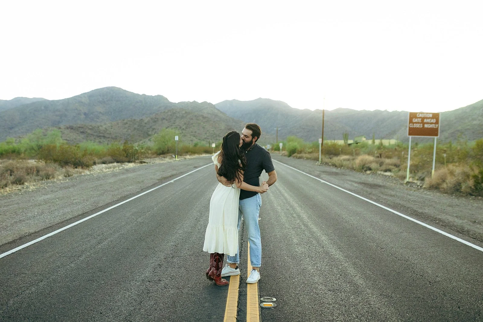 A couple kissing in the middle of a deserted road in a desert landscape with mountains in the background during daylight.