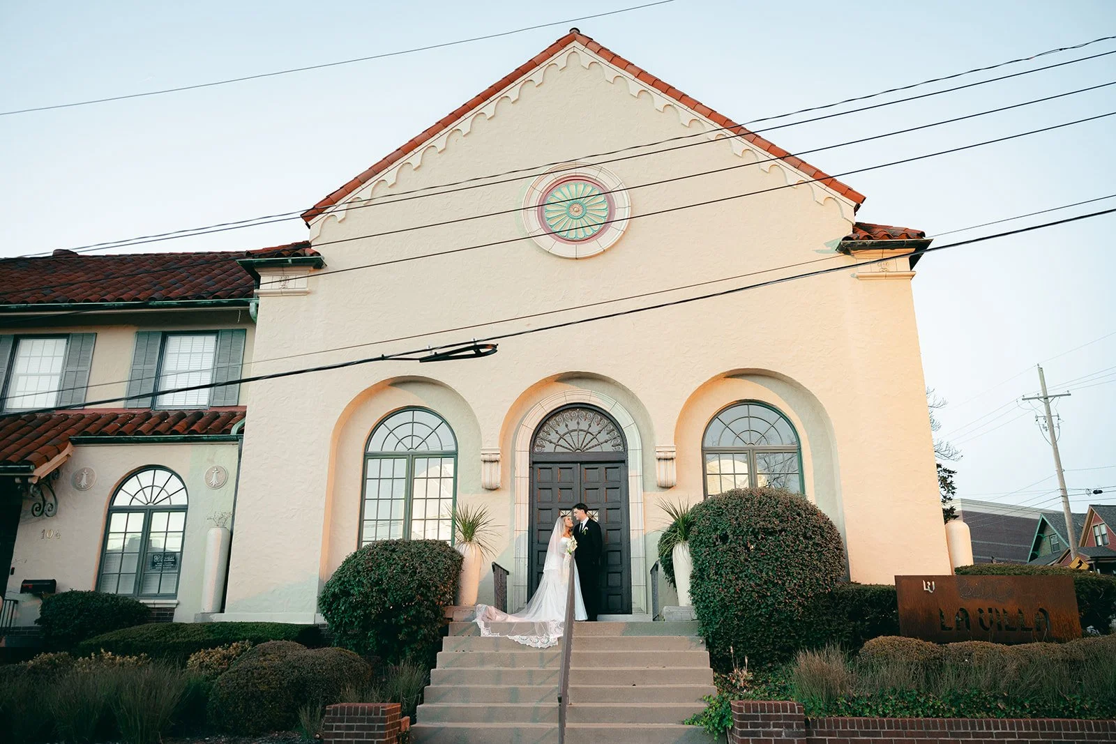 A bride and groom standing on the steps of a light-colored chapel, holding hands and looking at each other, with bushes and a sign that says "La Iglesia."
