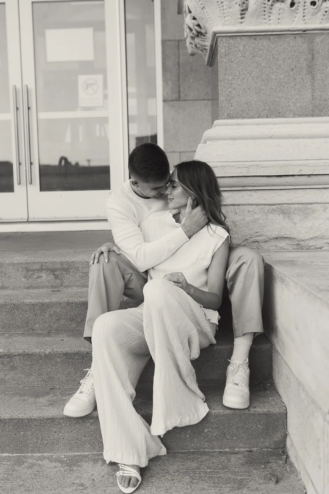 A man and woman sit on stairs outside a building, leaning in for a gentle kiss, with the man's hand around the woman's neck. They are wearing casual clothing, and the woman has long hair and wears earrings.