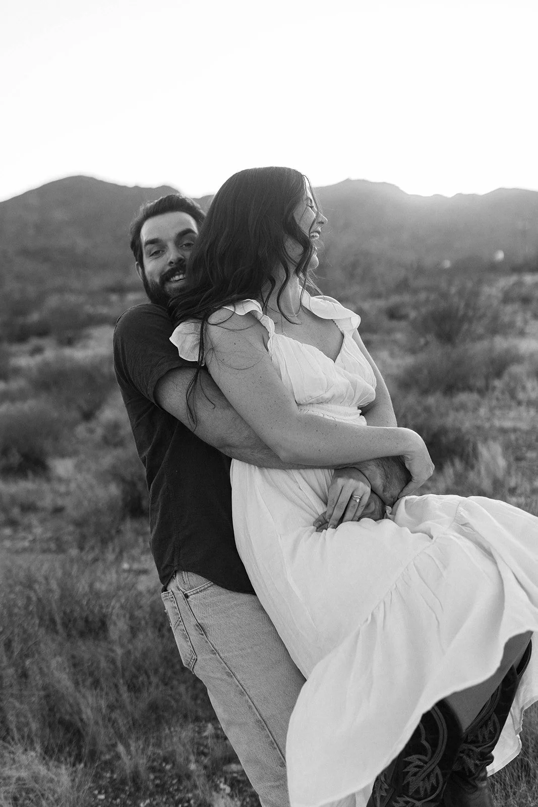 A black-and-white photo of a man lifting and holding a woman in an outdoor landscape with mountains in the background.