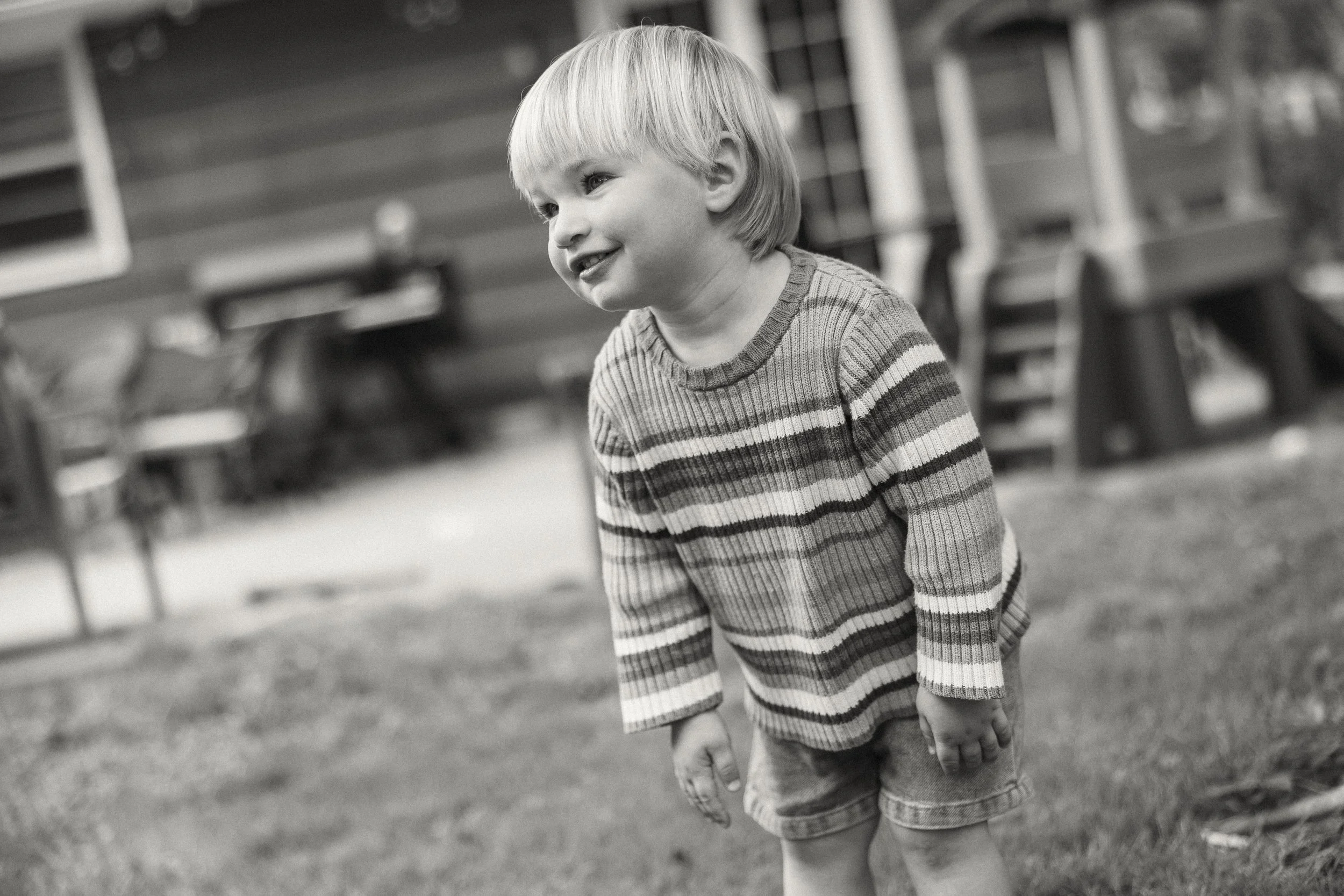A young boy with light hair and a striped sweater standing outdoors near a row of benches, smiling and looking to the side.