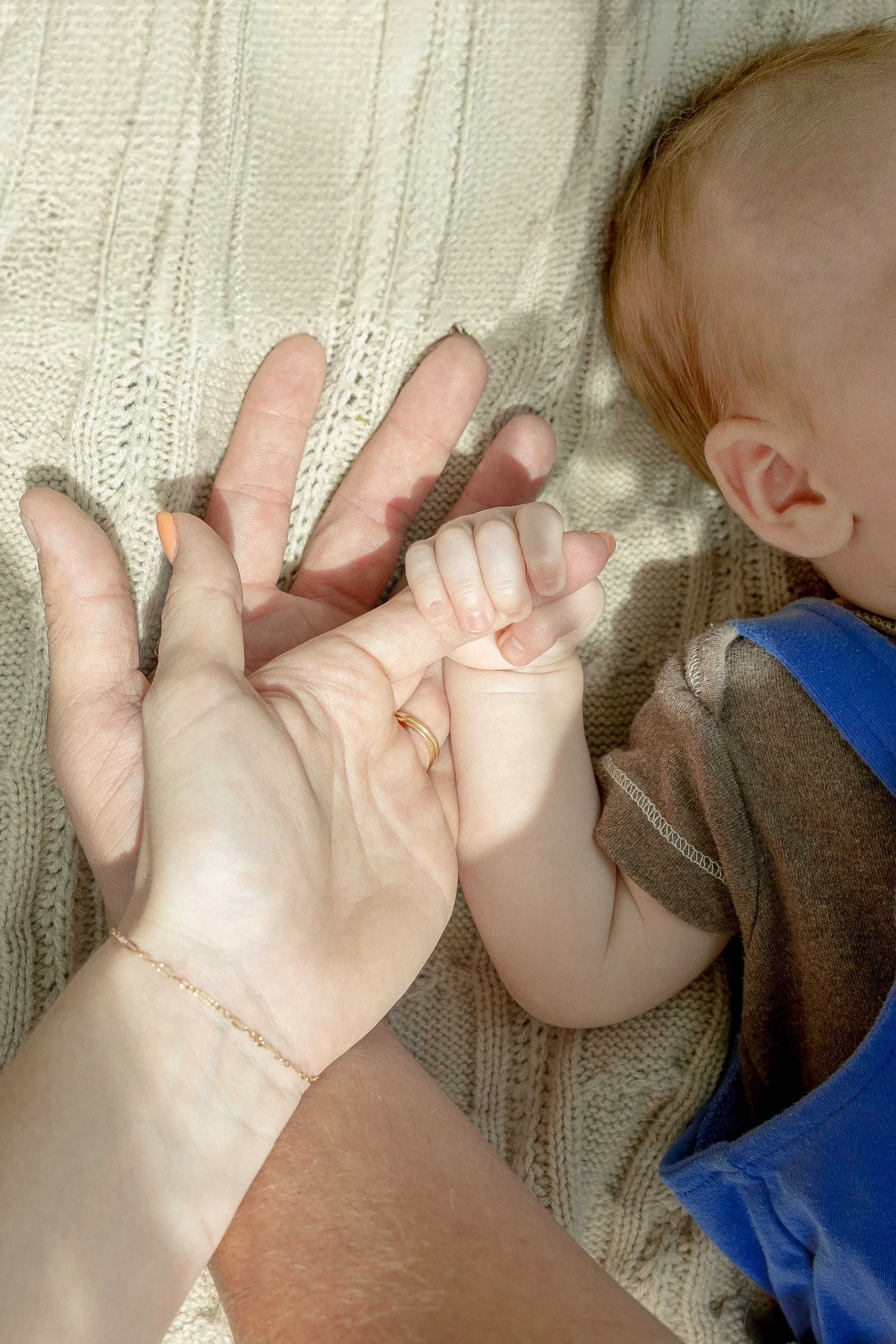 A hand and a child's hand holding together on a textured beige blanket, with the child's head resting nearby.