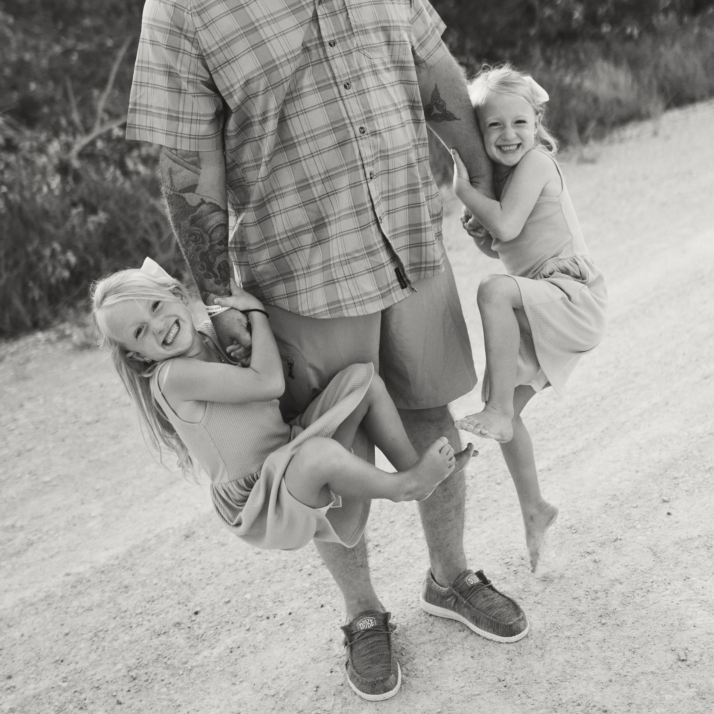 A black and white photo of two young girls hugging an adult man, with the girls smiling and holding hands with him. The girl on the left is hugging the man's leg, and the girl on the right is hugging his arm. They are outdoors on a dirt path with trees in the background.