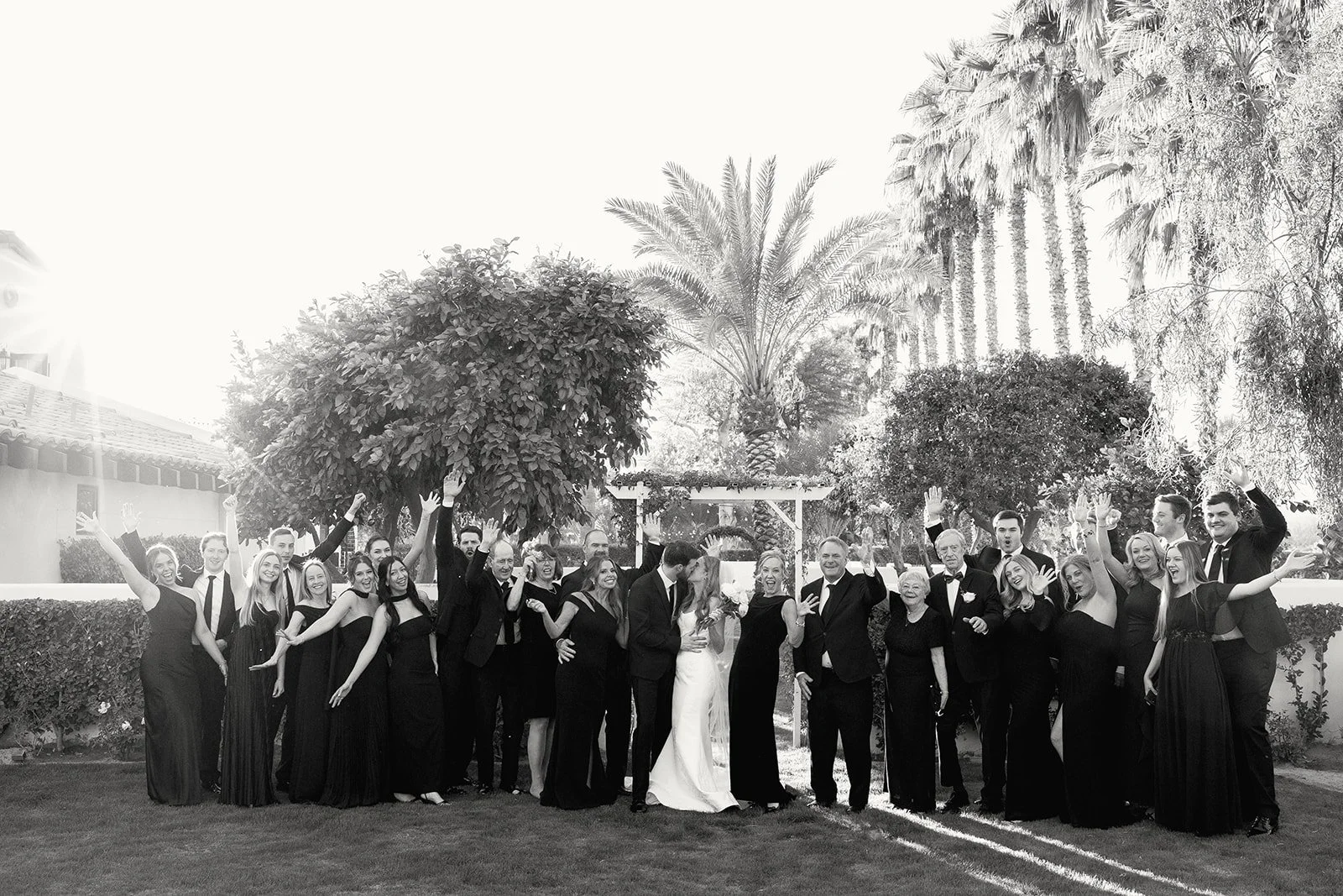 A group of people in formal attire celebrating outdoors, with some raising their hands, trees, and a clear sky in the background.