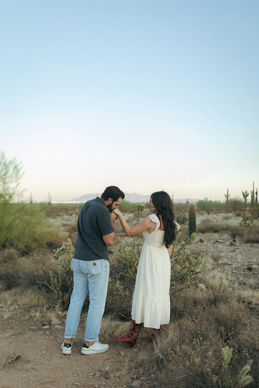 A man and woman holding hands in a desert landscape with cacti and mountains in the background, she is wearing a white dress and cowboy boots, he is in a dark shirt and jeans.