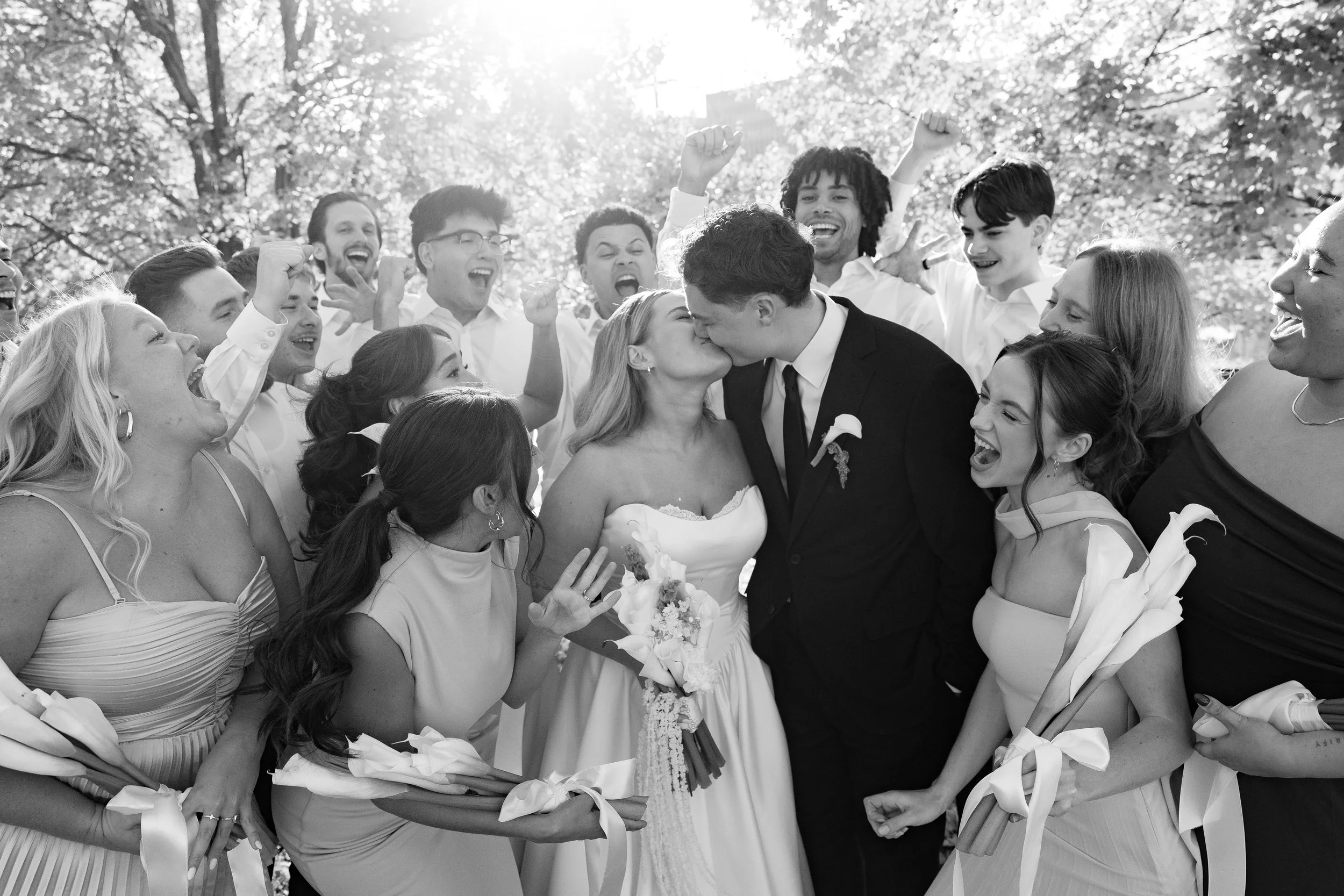A black and white photo of a wedding celebration with a bride and groom kissing, surrounded by smiling guests holding bouquets and raising their hands in excitement, outdoors with trees and sunlight in the background.