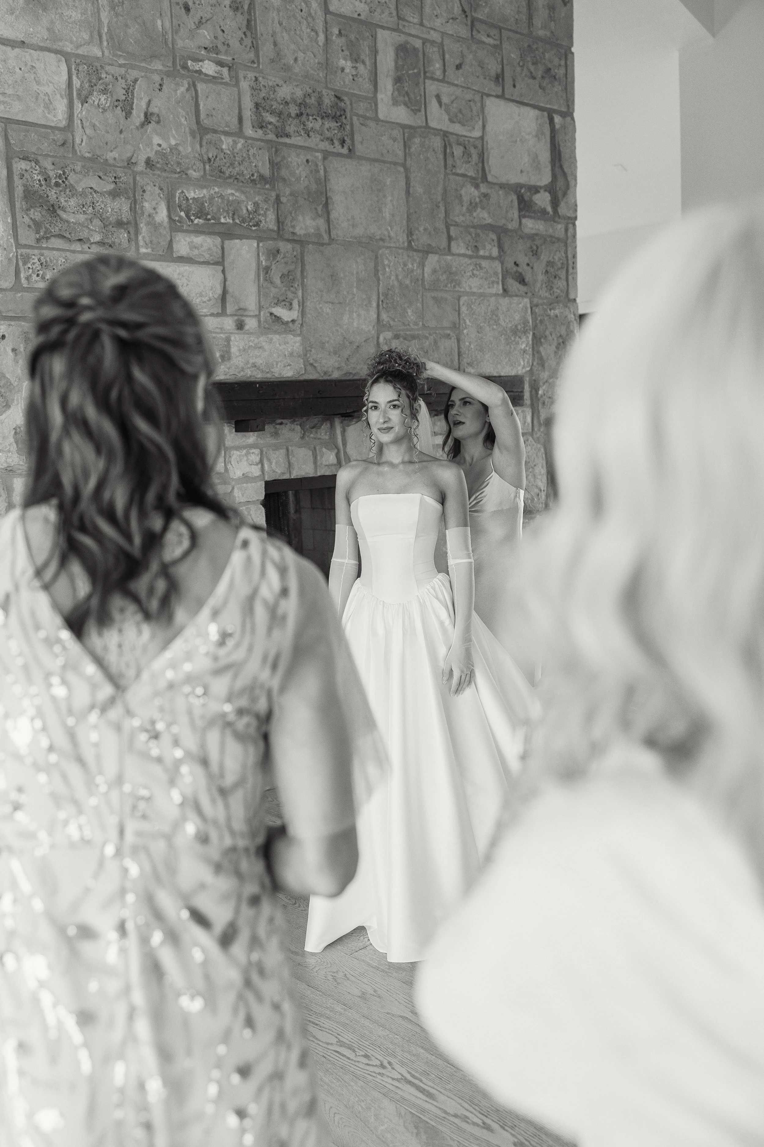 A bride in a strapless wedding gown and long gloves stands near a stone fireplace, with a woman fixing her hair, while multiple women observe in the foreground, in an indoor setting.