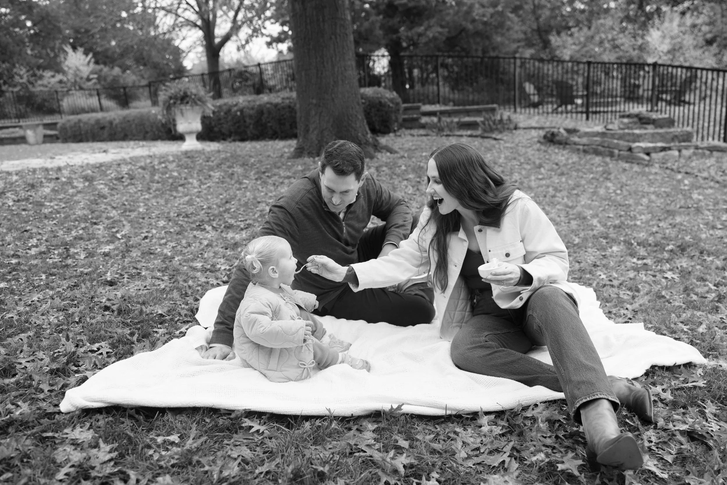 A family of three, including a man, a woman, and a young girl, sitting on a blanket in a park during fall. The woman is feeding the girl while the man watches and smiles. Fall leaves are scattered on the ground, with trees and a fenced area in the ba