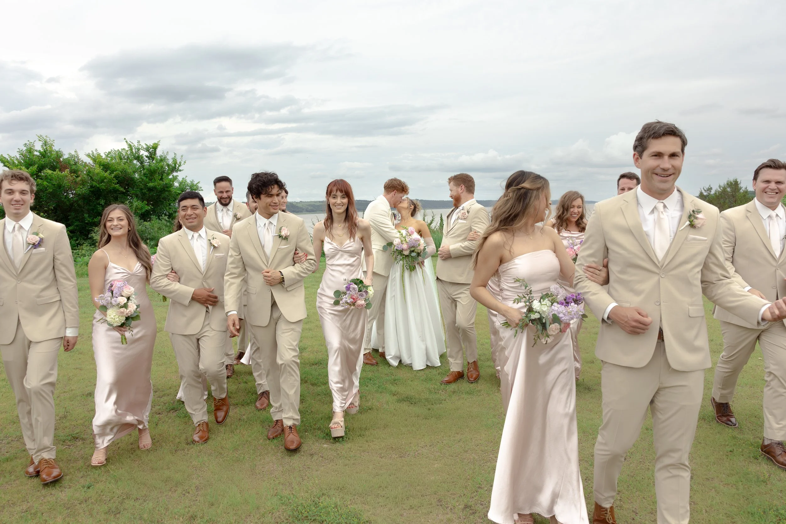 A bridal party walking outdoors on a grassy field during a wedding celebration. Some are holding bouquets of flowers, and the bride and groom are in the background kissing. Wedding in Manhattan, Kansas at Blue Vista in the Flint Hills. 