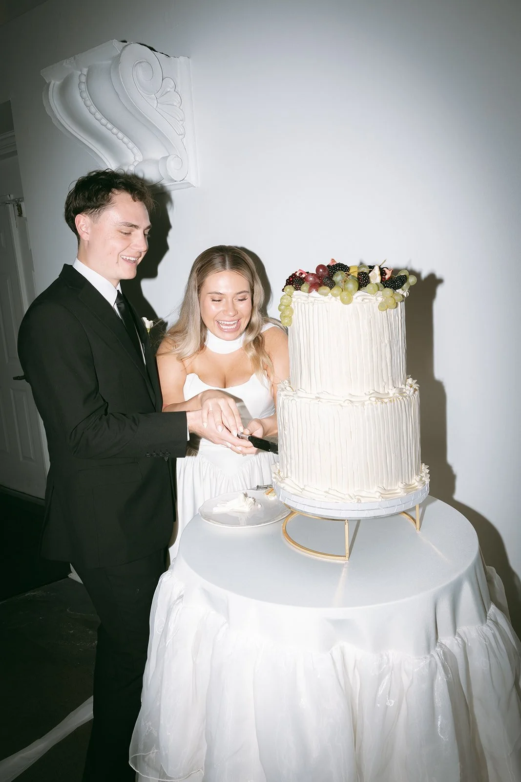 A newlywed couple cutting a wedding cake at their celebration, with a white, two-tier cake topped with assorted fresh grapes and berries, in a decorated venue.