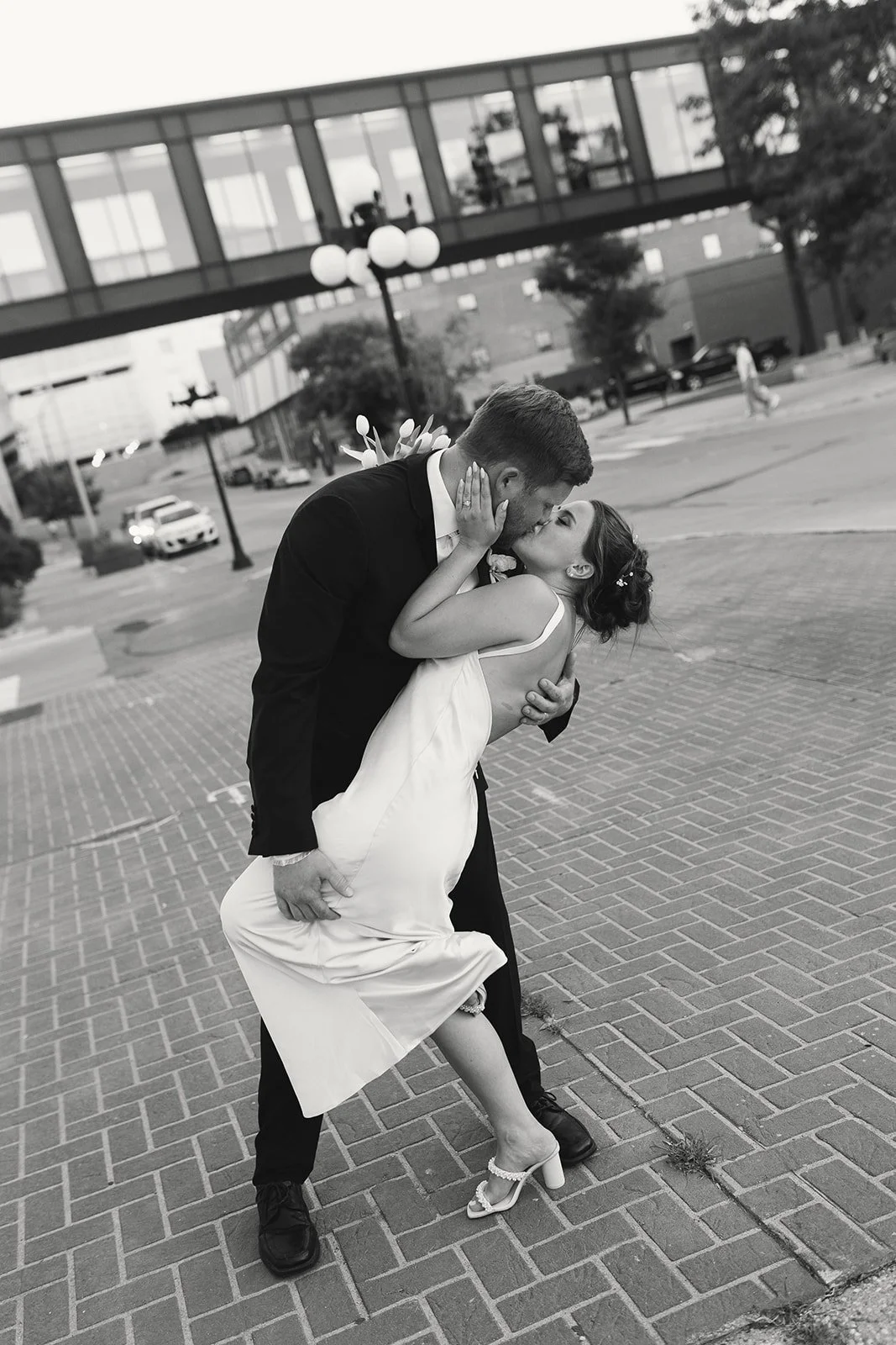 A couple, dressed in wedding attire, sharing a passionate kiss outdoors on a city street, with the groom bending over the bride who is lifting one leg.