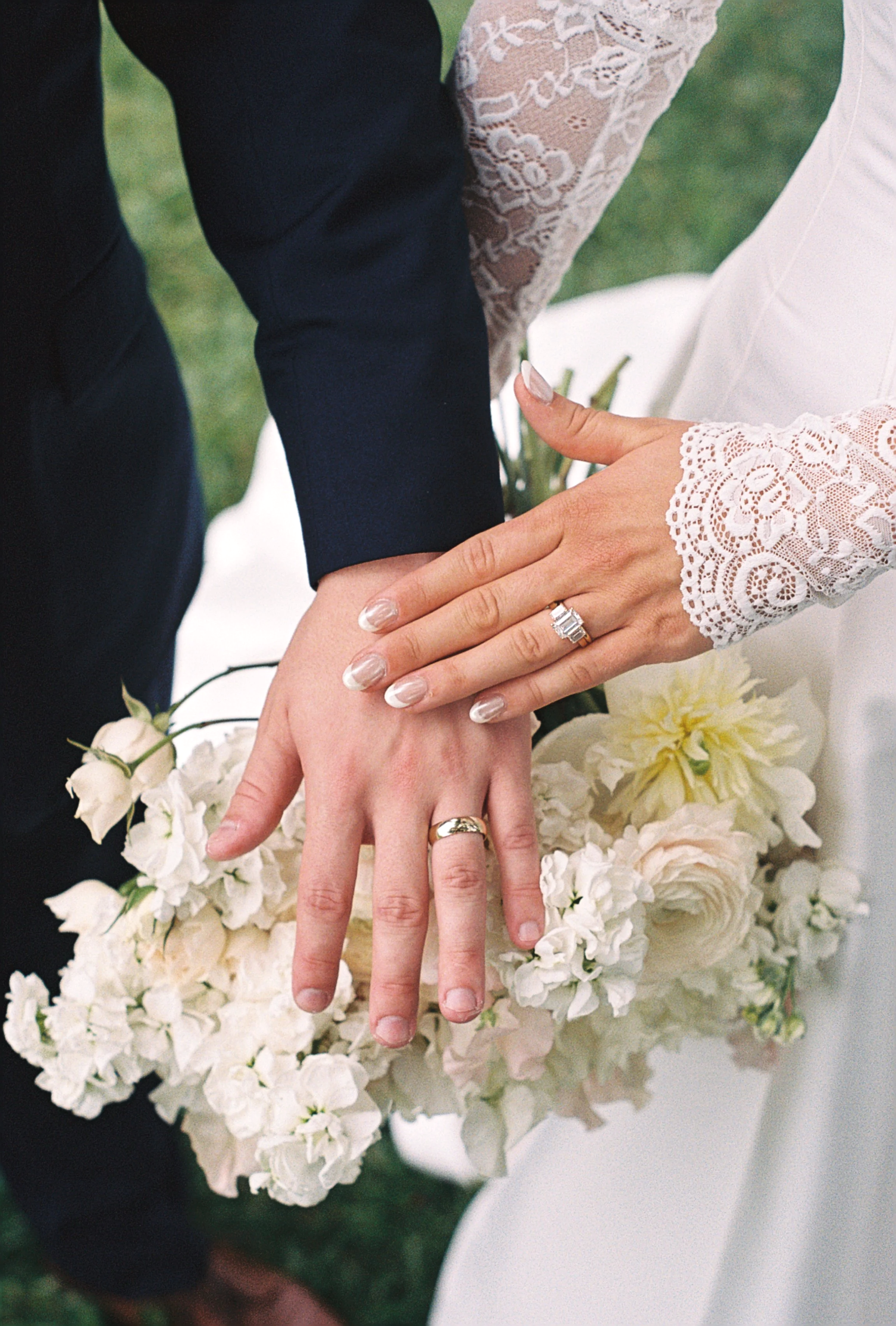 Close-up of a couple's hands showing wedding rings with a bouquet of white flowers in the background. The bride is wearing a lace sleeve.