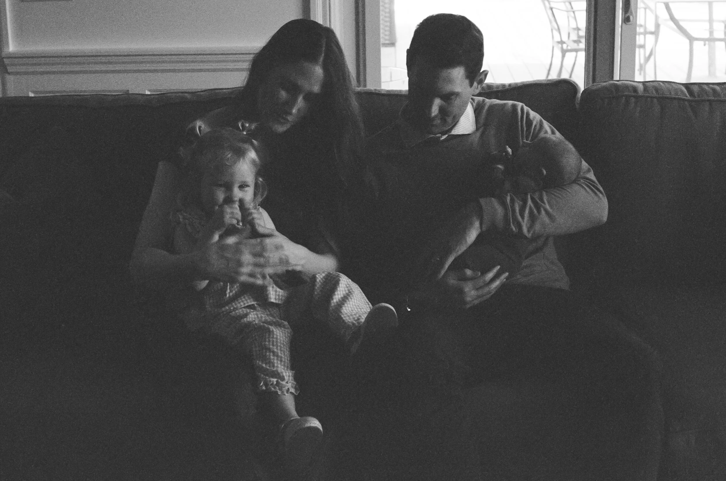 A black-and-white photo of a family sitting on a sofa in a living room near a sliding glass door. A woman, a man, and a young girl are visible, with the girl smiling and sitting with her legs crossed. The woman and man are looking down at something t