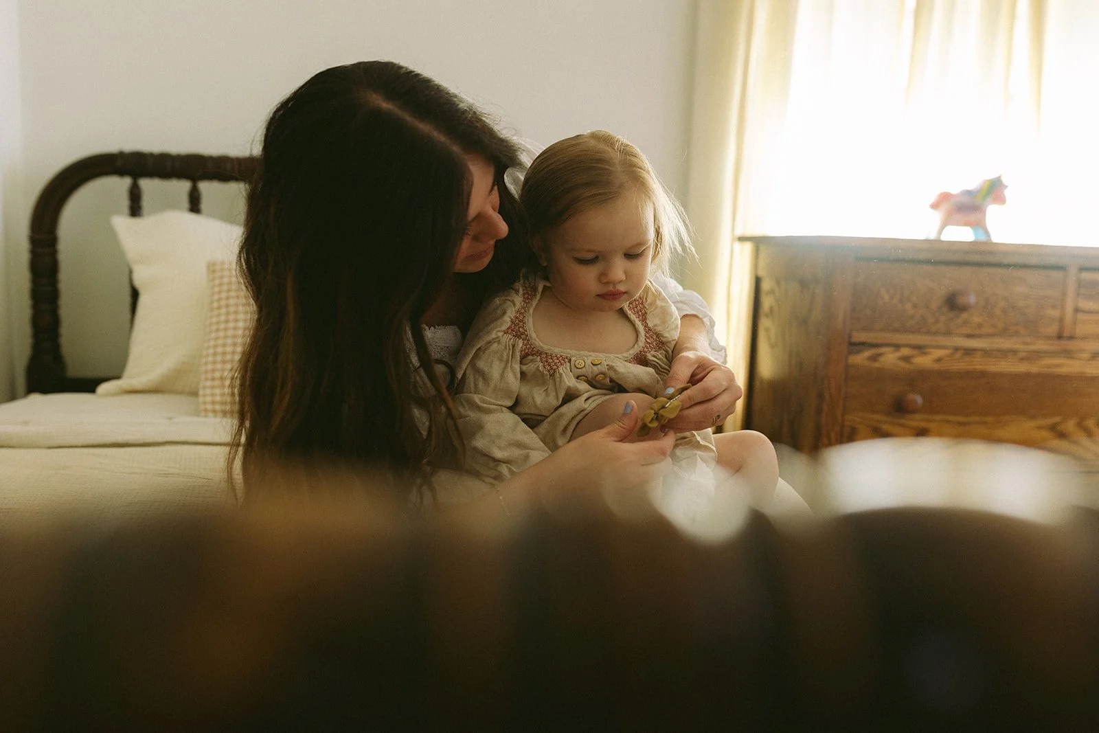 A woman and a young girl sit on a bed, looking at a small object in the woman's hands. The room has a wooden dresser, a window with curtain, and a toy on the dresser.