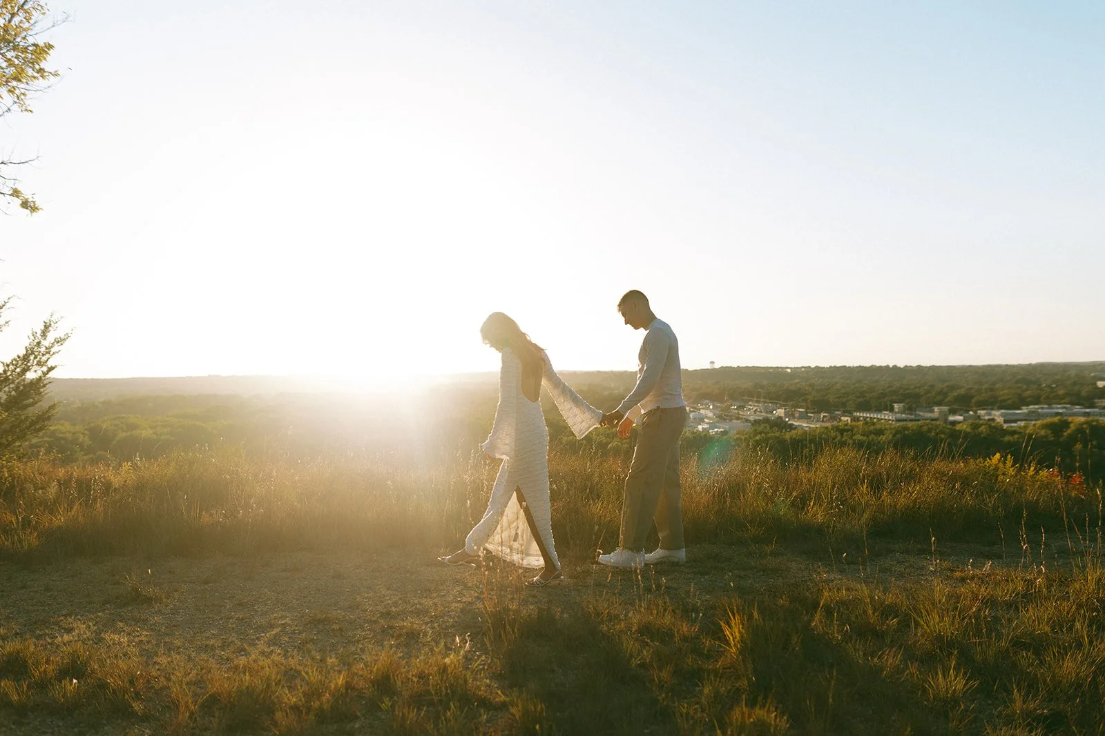 A couple holding hands walks through a grassy field during sunset.