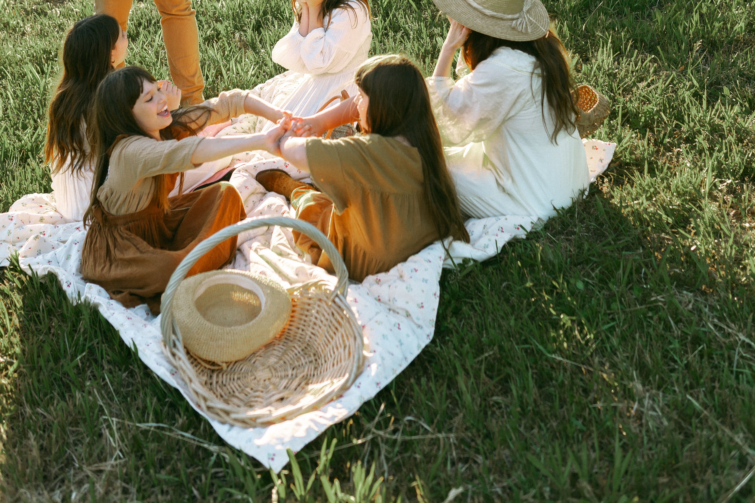 A group of women and girls having a picnic outdoors on a blanket in a grassy field, playing a tug-of-war game with a rope, with a straw hat on a basket in the foreground.