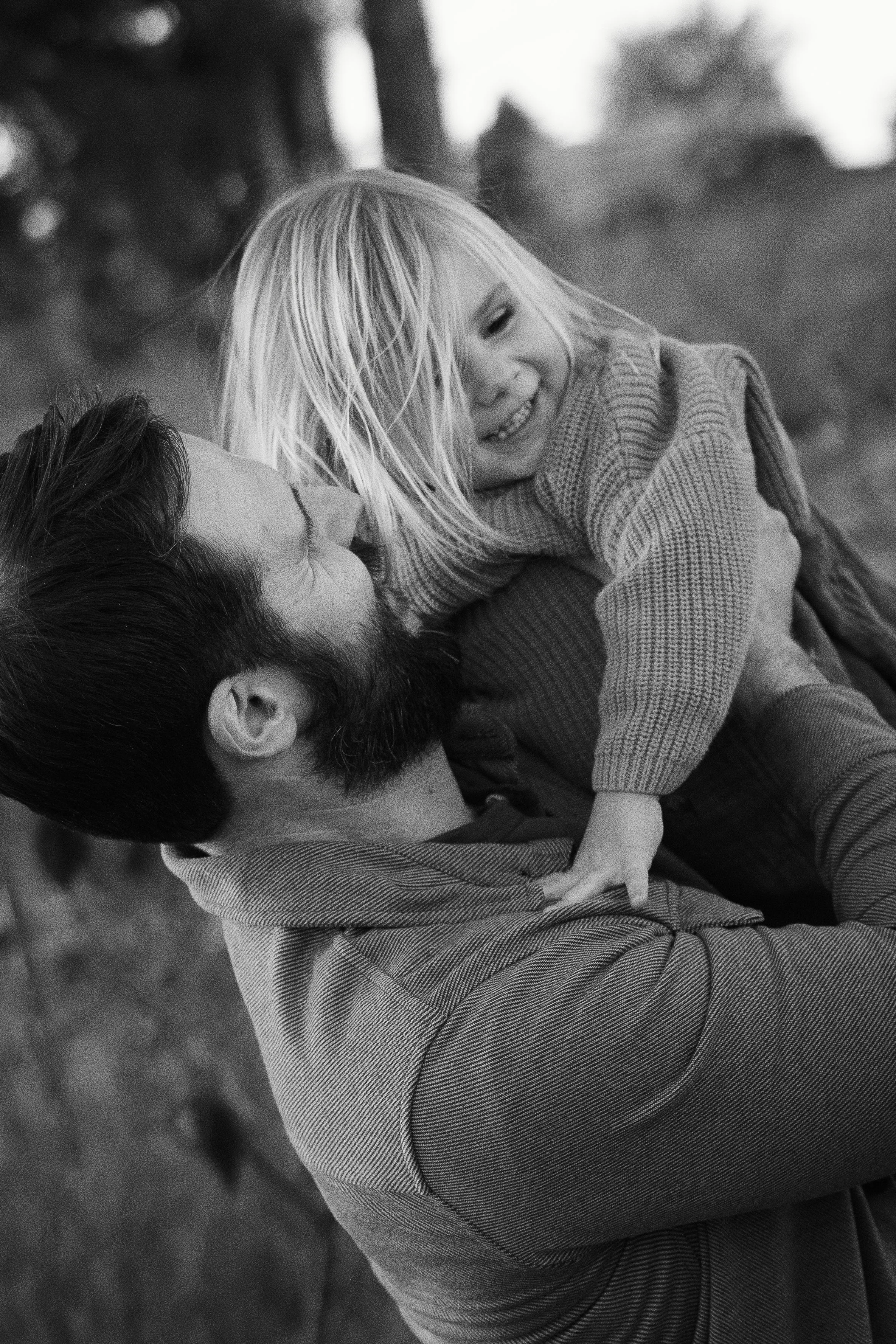 A man with a beard holding a young girl with long blonde hair in his arms, both smiling and enjoying a moment outdoors, black and white photo.