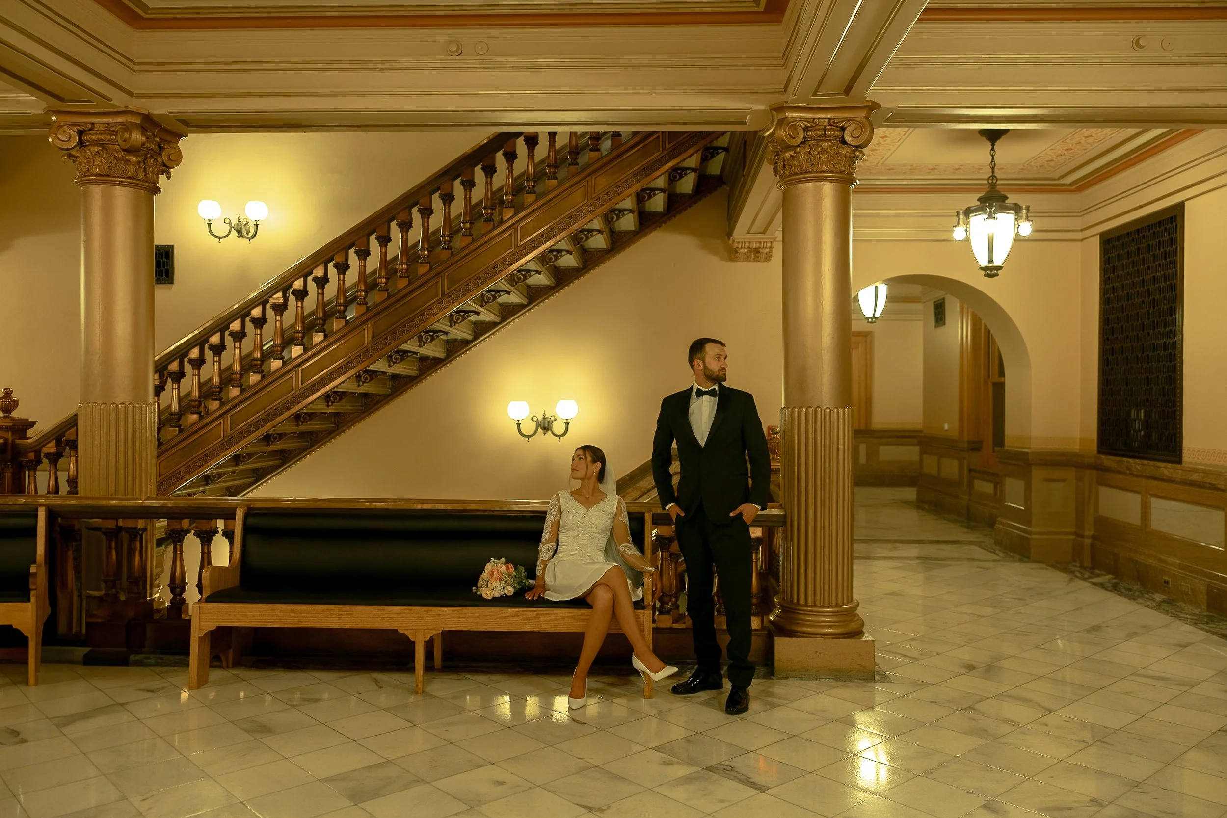 A bride and groom stand in a luxurious, ornate hotel lobby. The bride is sitting on a bench, wearing a white wedding dress and high heels, with a bouquet of flowers beside her. The groom is standing next to her, dressed in a black tuxedo with a bow t