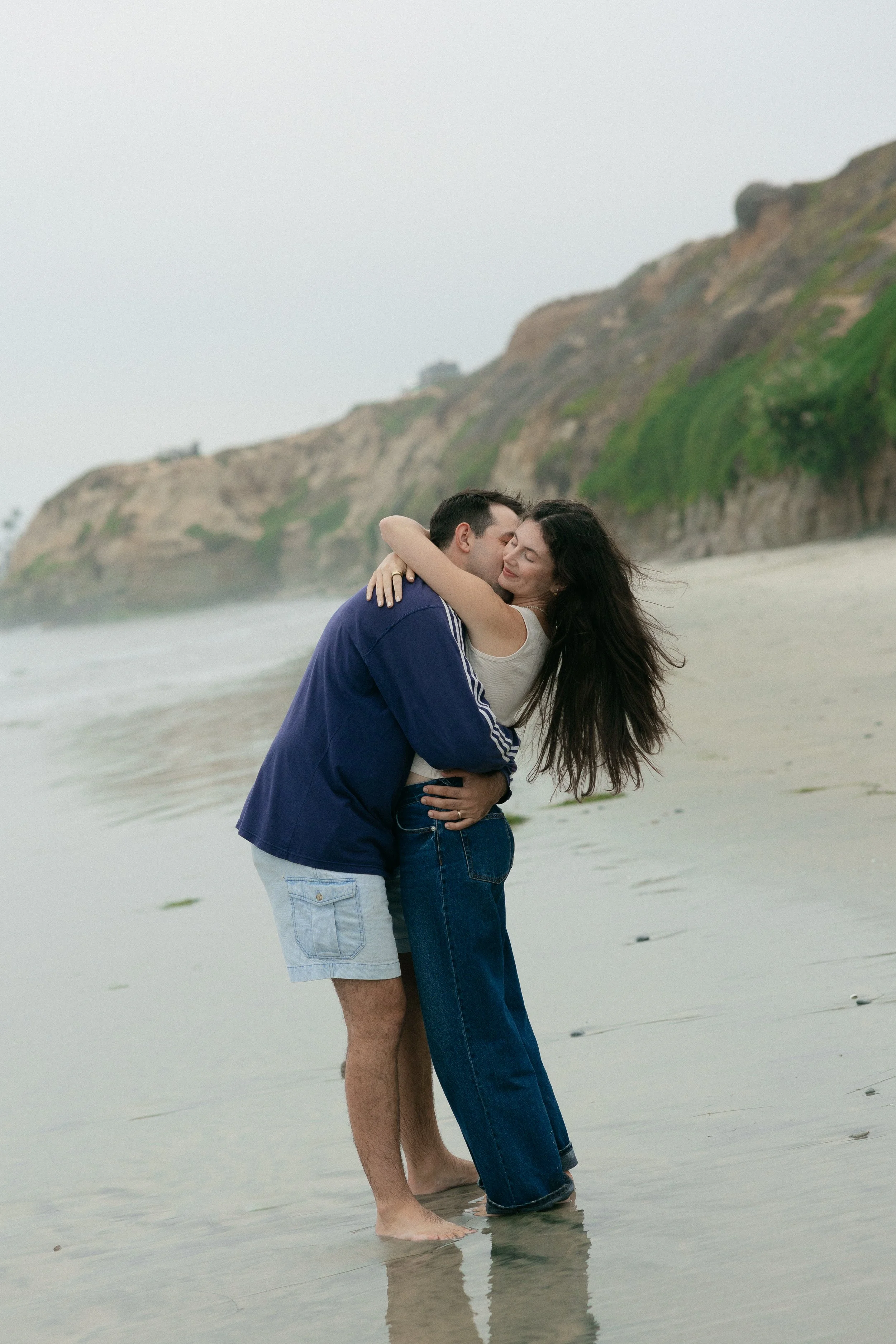 A couple hugging on a beach with cliffs in the background.