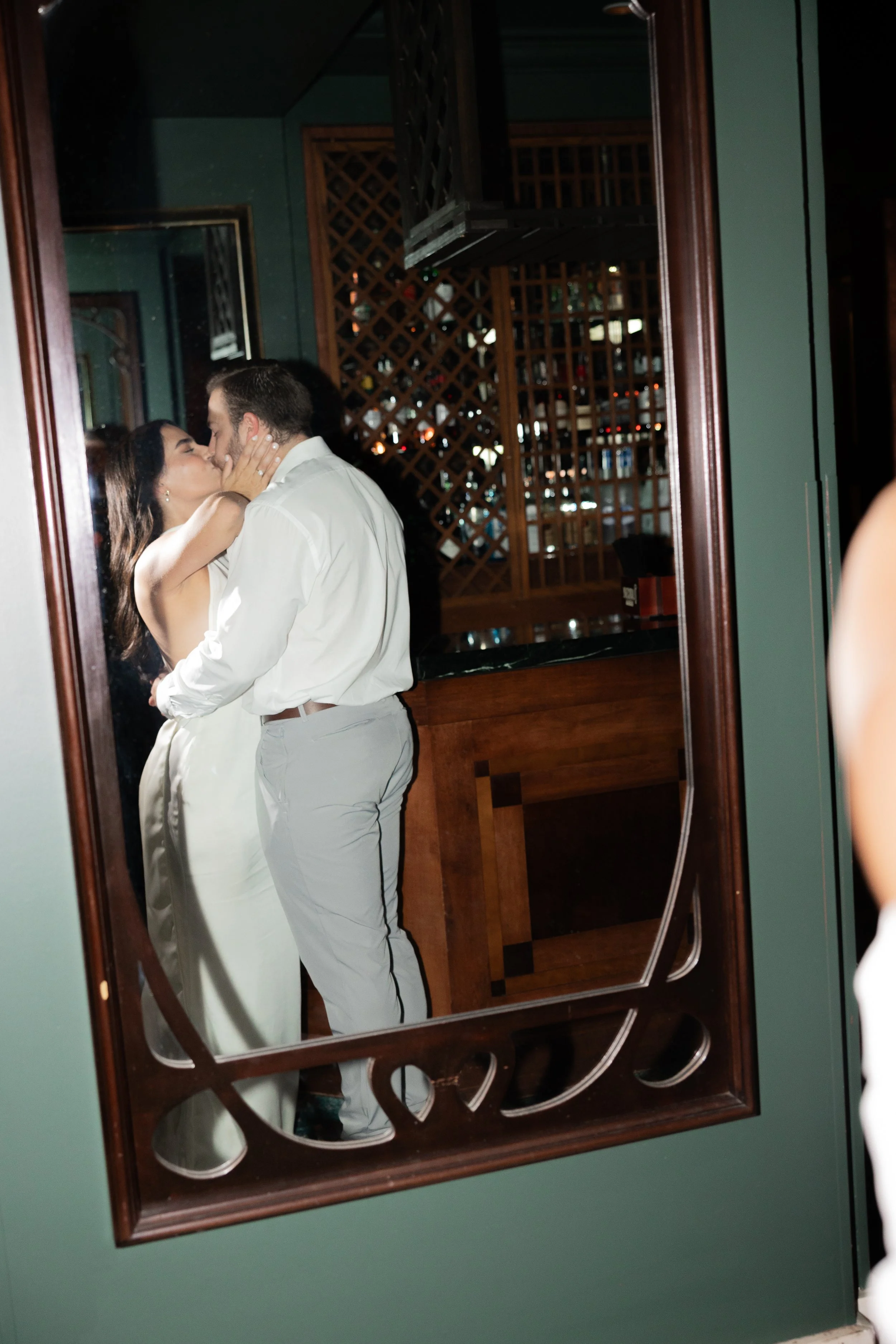 A couple kissing and embracing in front of a mirror, with a bar and wooden lattice in the background.