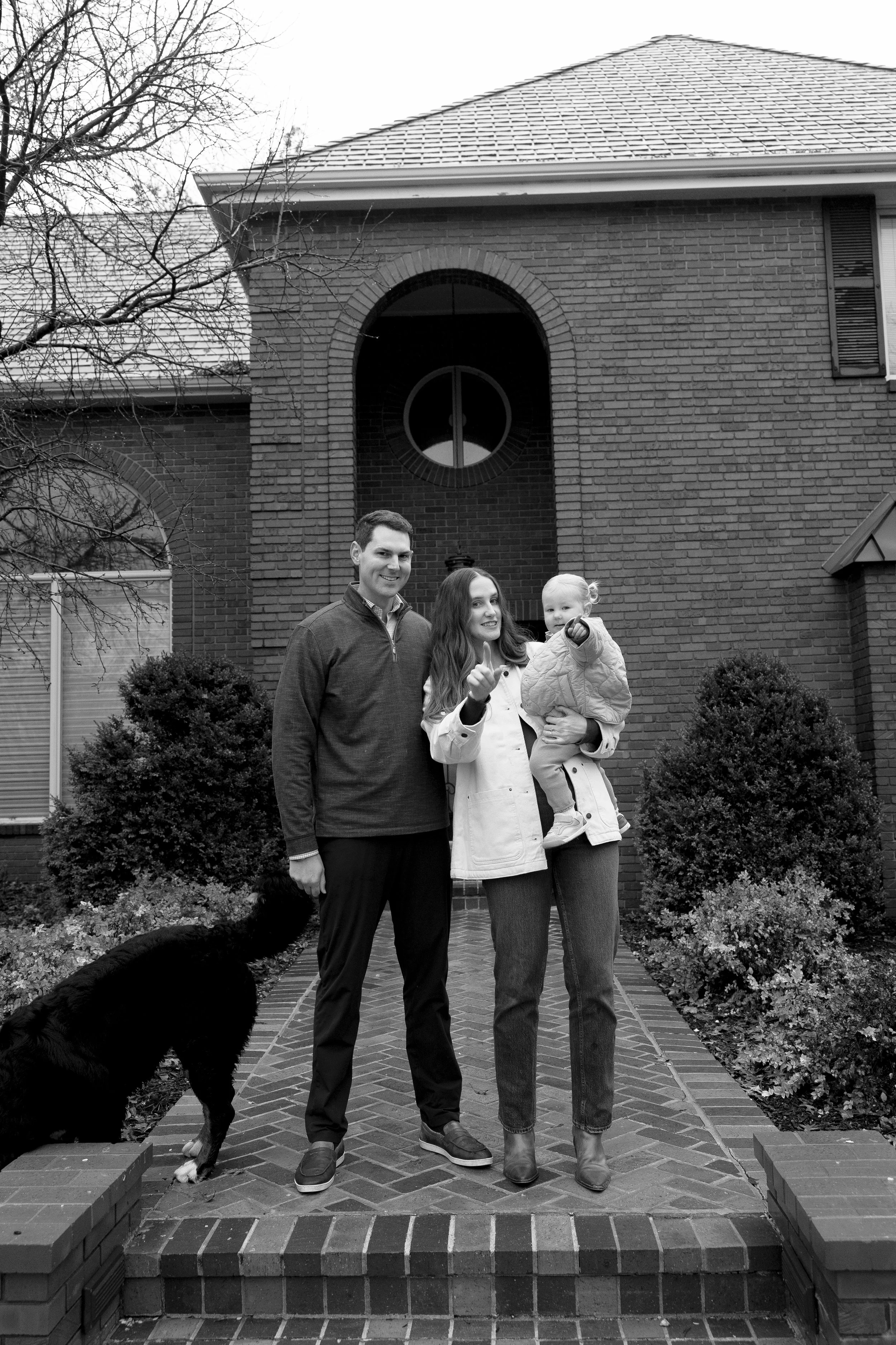 A family of three, with a dog, standing on their front porch in front of a brick house, smiling and making gestures.