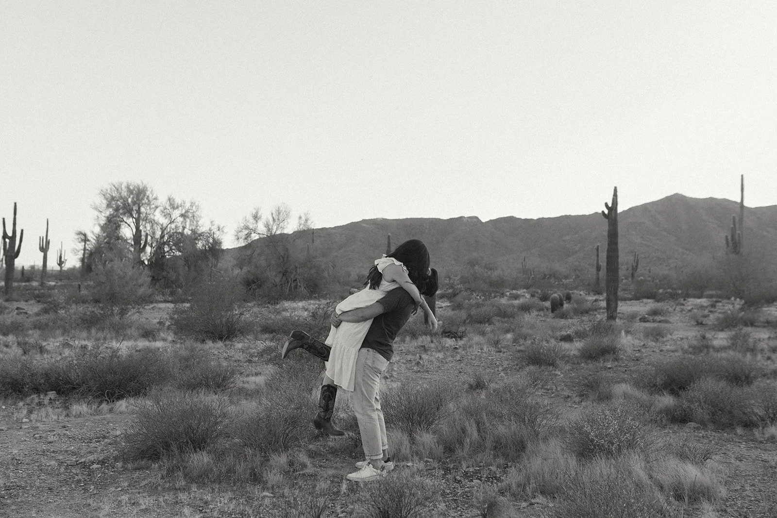 A person giving a piggyback ride to a child in a desert landscape with cacti and mountains in the background, in black and white.