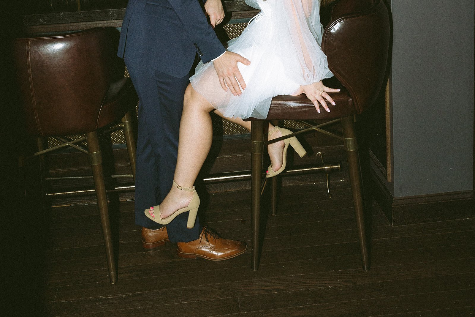 A person in a dark suit and brown shoes is standing beside a woman in a white skirt and beige high heels, who is sitting on a barstool with her legs crossed, showing her legs and feet.